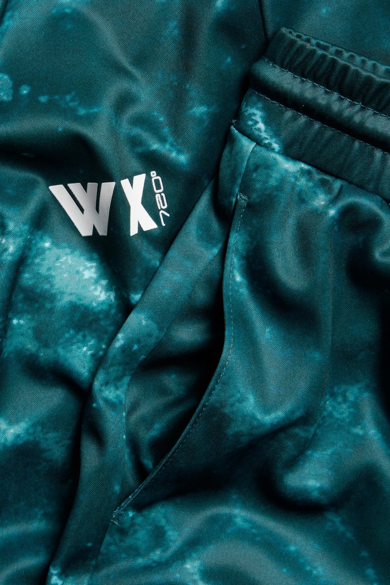 Zbliżenie zielonych, wzorzystych szortów sportowych z poliestru z kieszenią, elastycznym pasem i białym logo.