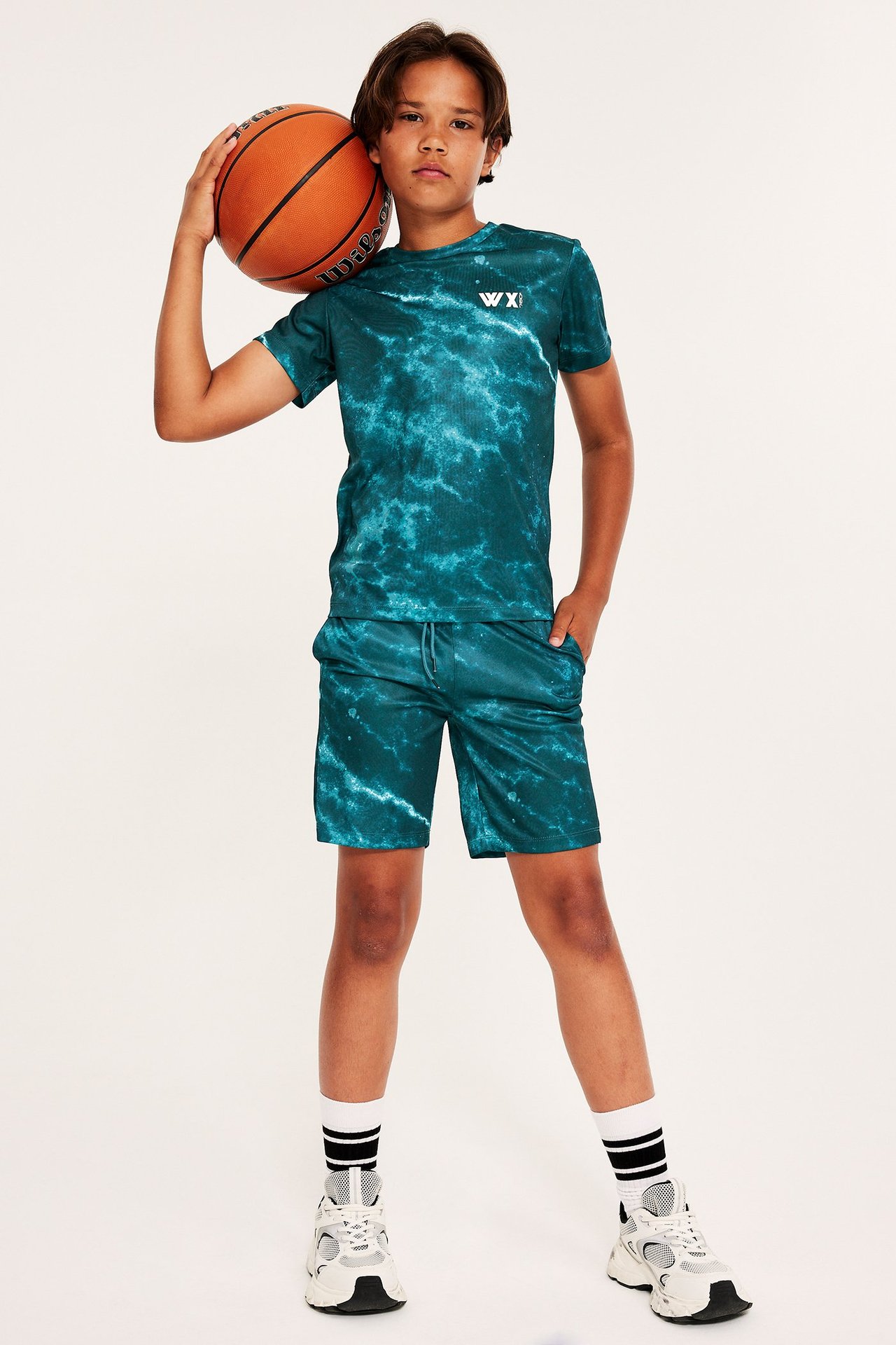 Widok z przodu: dziecko w zielonym wzorzystym T-shircie i pasujących szortach, trzymające piłkę do koszykówki.