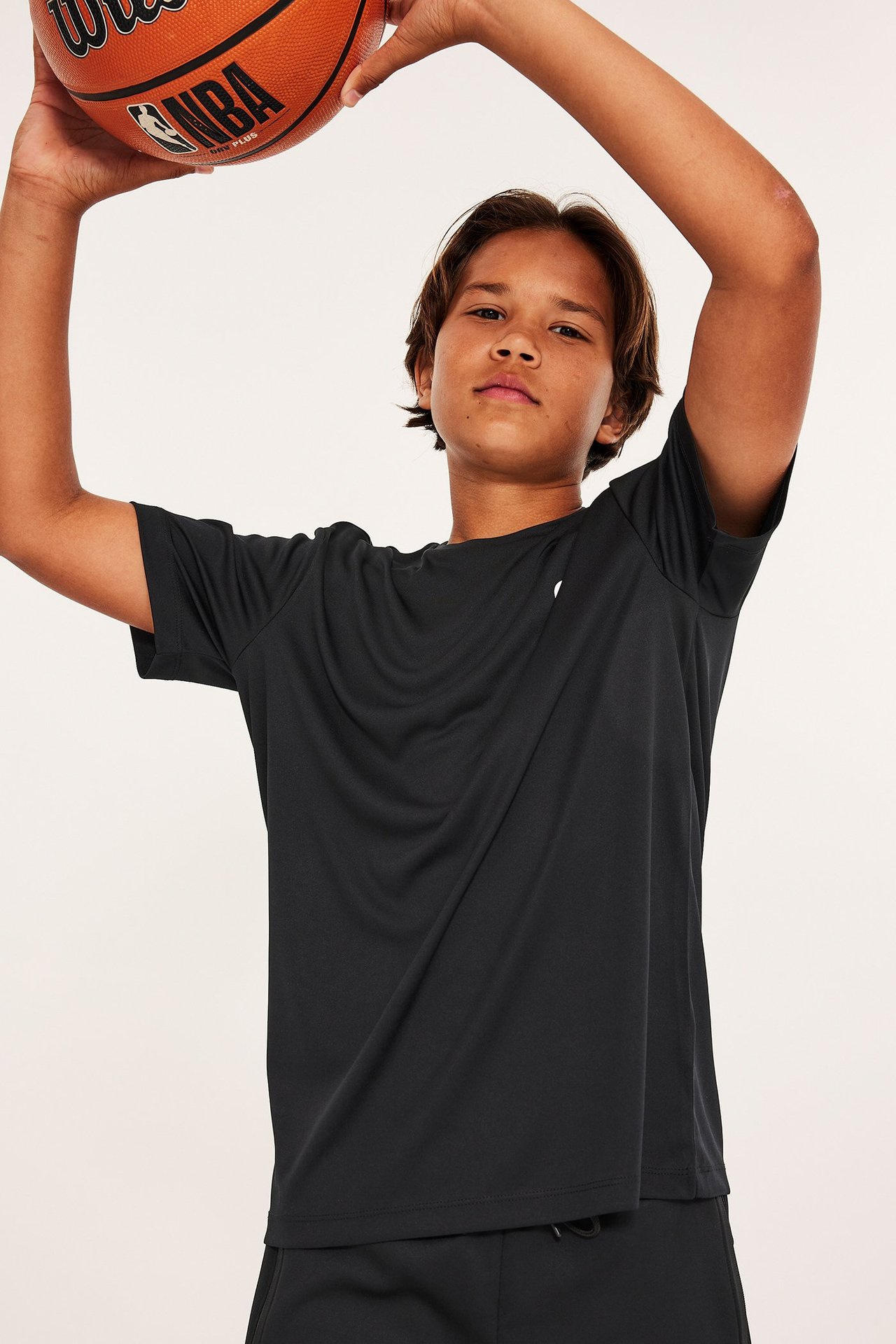 Widok z przodu dziecka w czarnej koszulce treningowej z poliestru z białym logo, trzymającego piłkę do koszykówki.