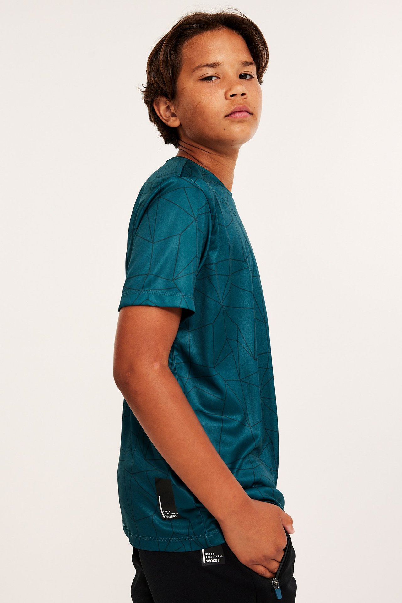 Widok z boku dziecka w ciemnoturkusowej, wzorzystej koszulce sportowej z poliestru i czarnych szortach.