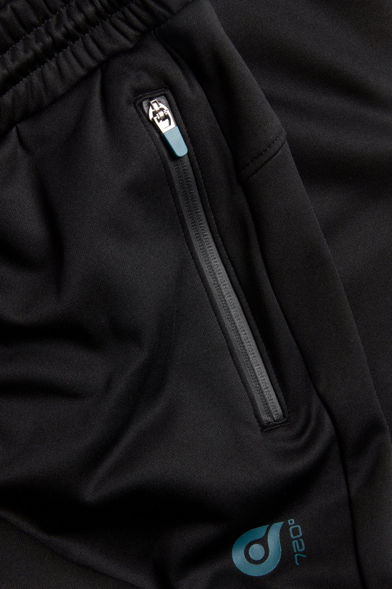 Zbliżenie czarnych poliestrowych spodni dresowych z elastyczną talią, kieszenią na zamek i turkusowym logo.