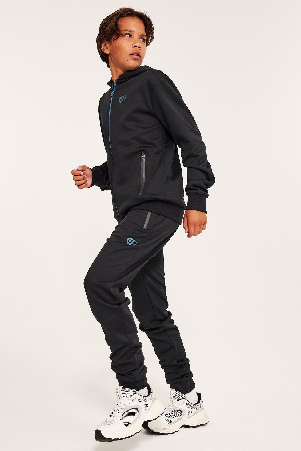 Widok z boku dziecka w czarnych poliestrowych spodniach dresowych z elastycznymi mankietami, czarnej bluzie z kapturem i białych sneakersach.