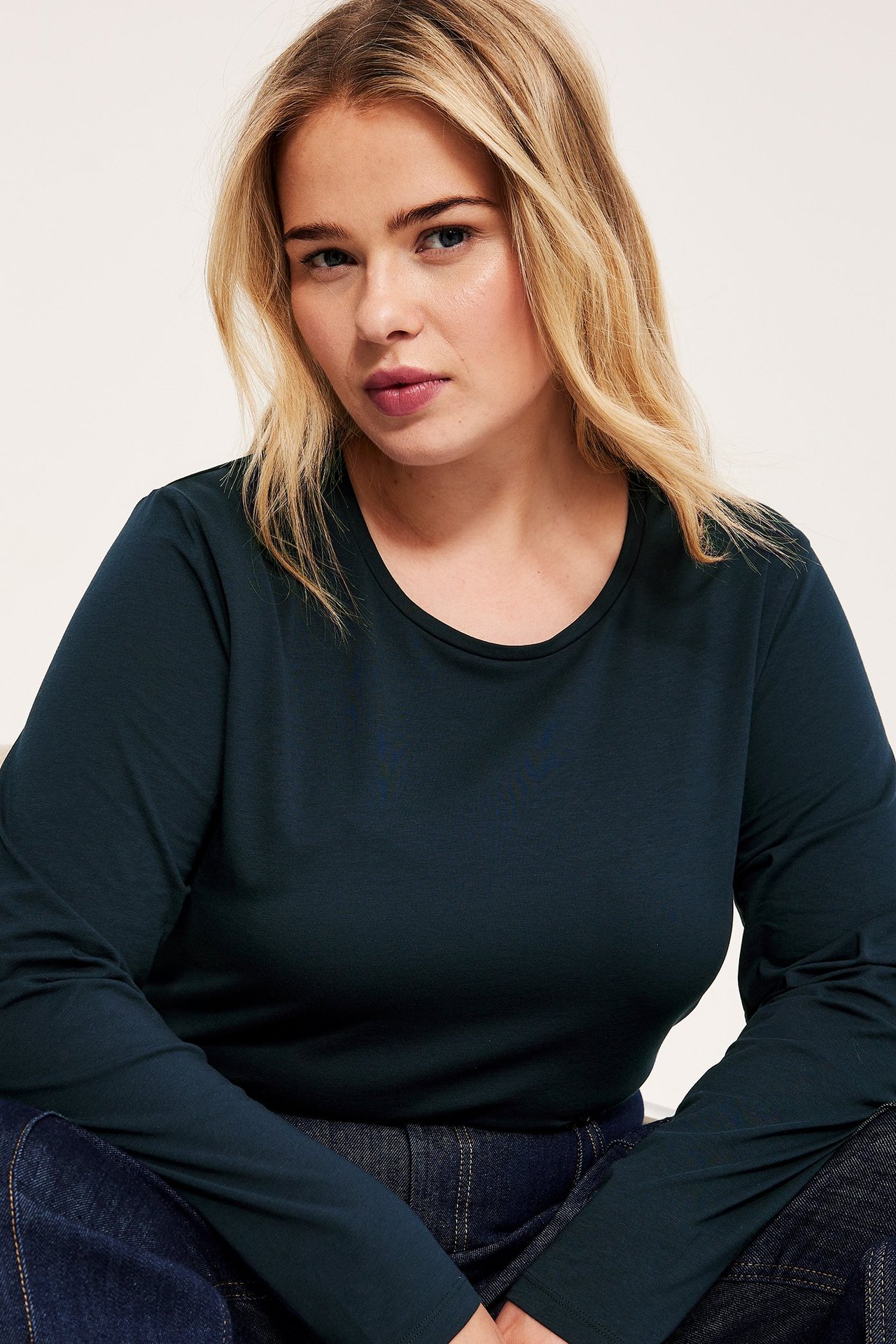 Nærbilde av en kvinne i en mørkeblå, ensfarget, langermet, elastisk bomulls-basistopp og mørkeblå jeans.