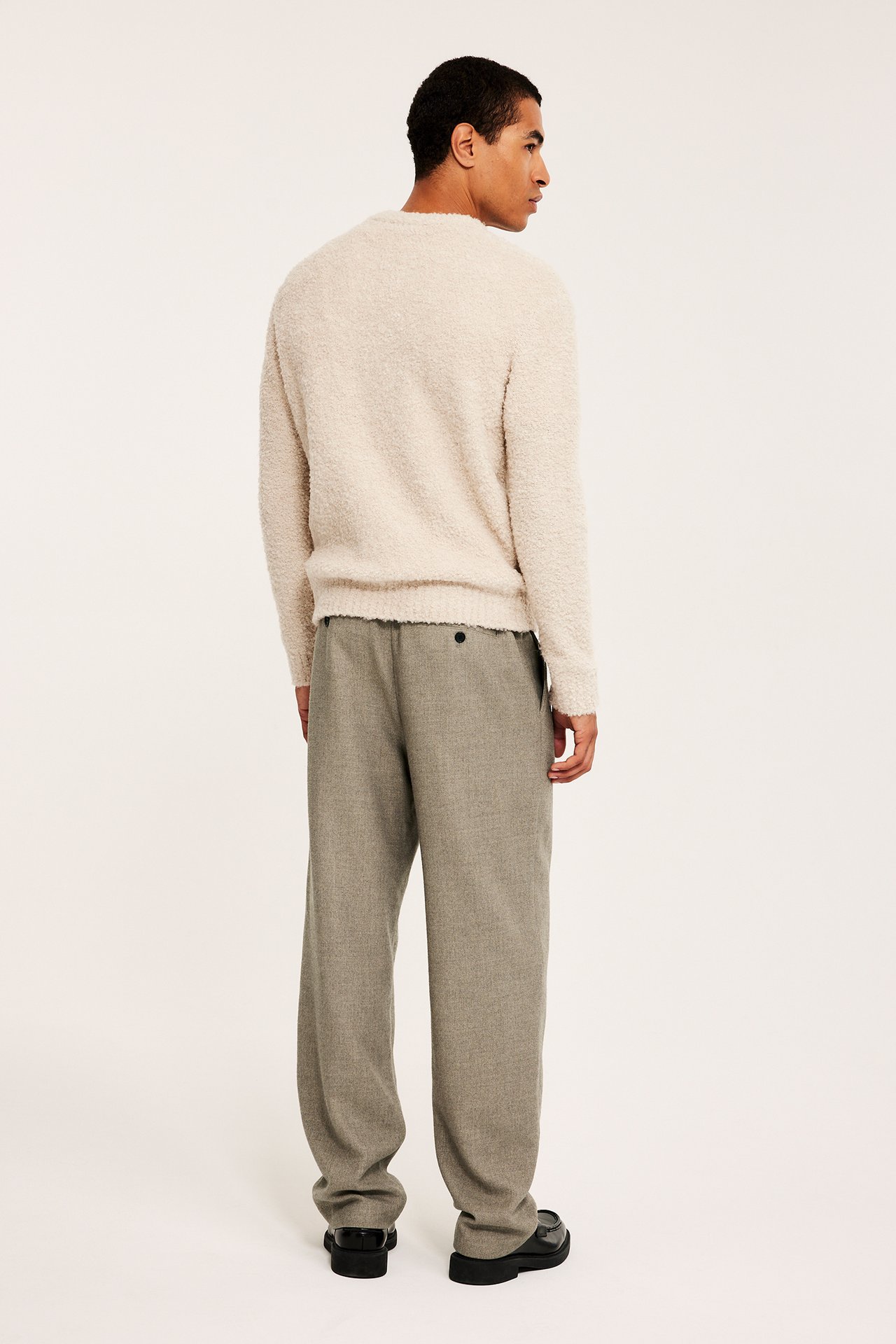 Bakifrån: Man i lösa beige melange borstade byxor, en krämvit fluffig tröja och svarta loafers.