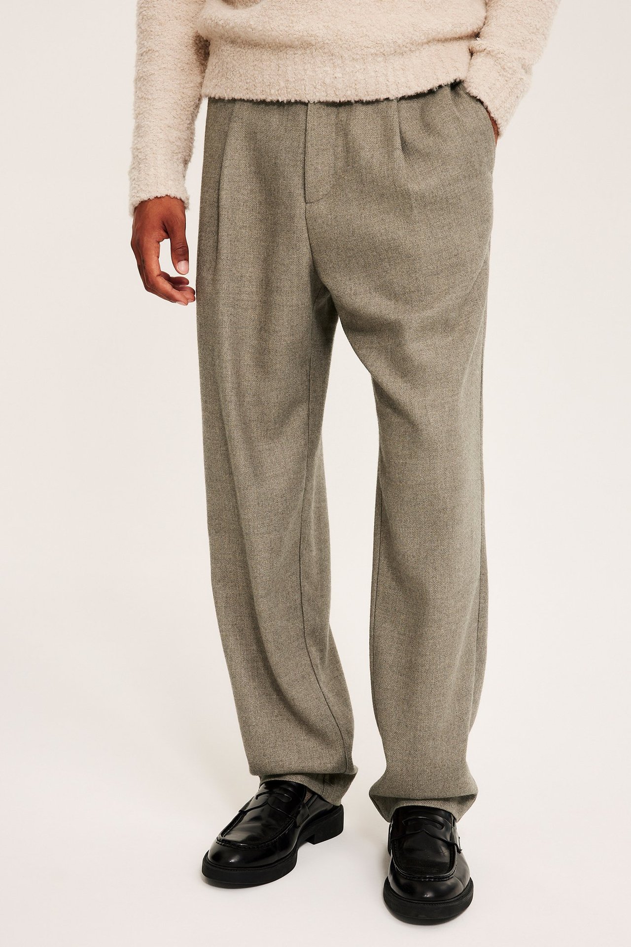 Framifrån av en man i beigemelange plisserade byxor, en beige fluffig tröja och svarta loafers.