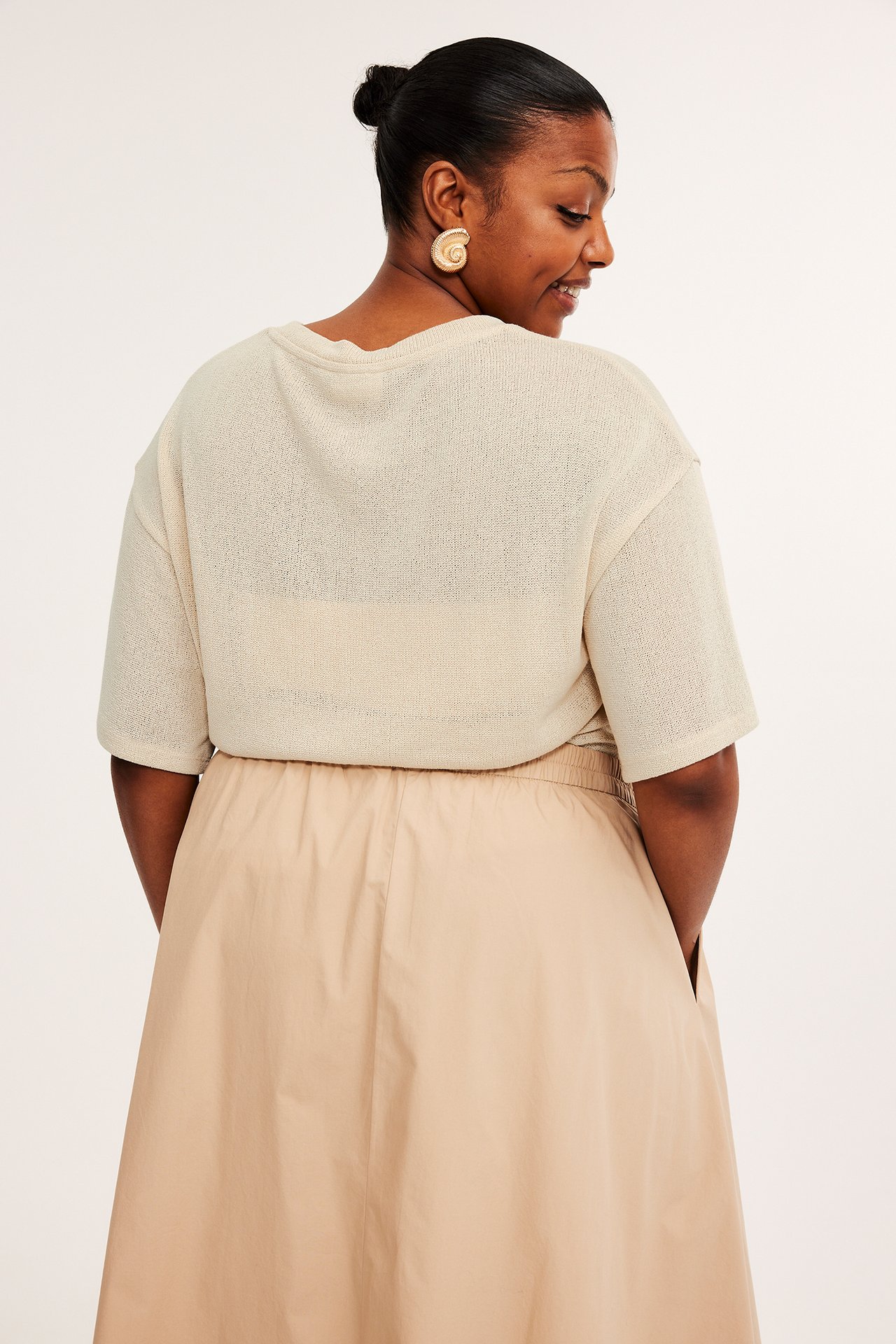 Bakifrån: kvinna i offwhite strukturerad polyester T-shirt, beige kjol och guldfärgade spiralörhängen.