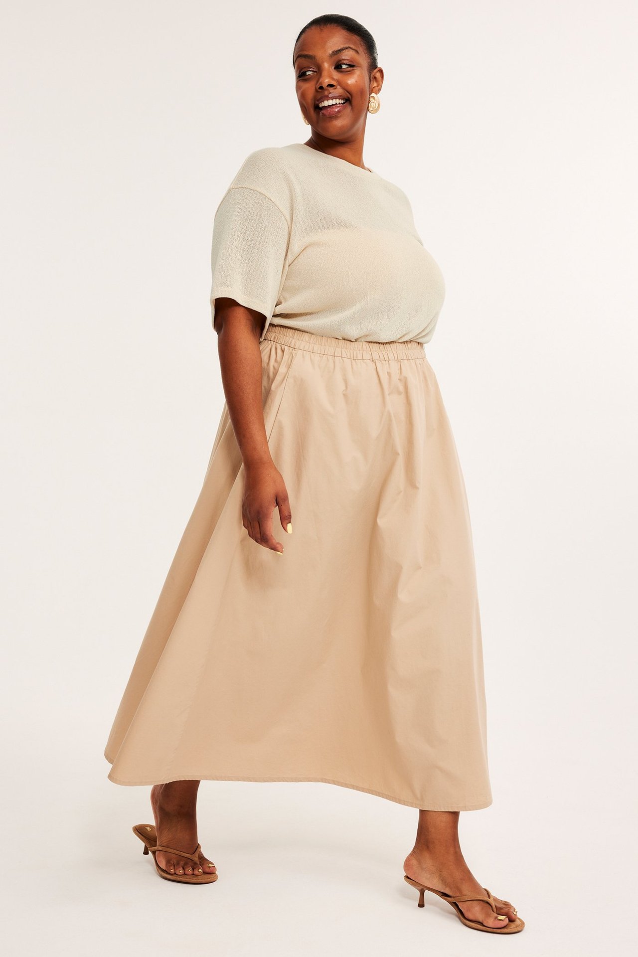 Forfra: kvinne i offwhite strukturert T-skjorte, beige midiskjørt, brune sandaler og gulløreringer.