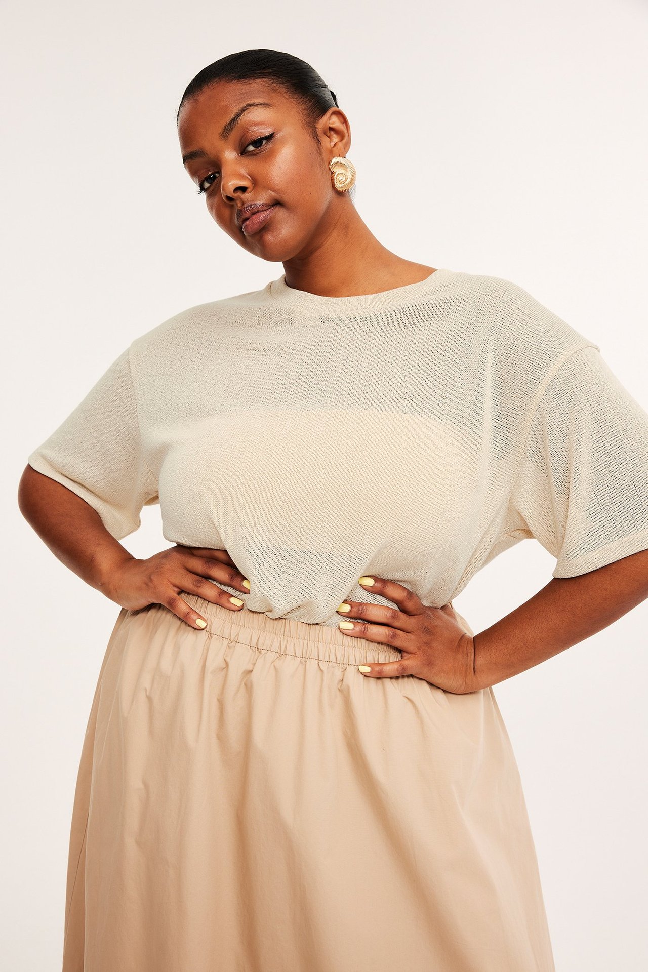 Widok z przodu: kobieta w kremowym teksturowanym t-shircie na jasnym topie, beżowej spódnicy i złotych kolczykach spiralnych.