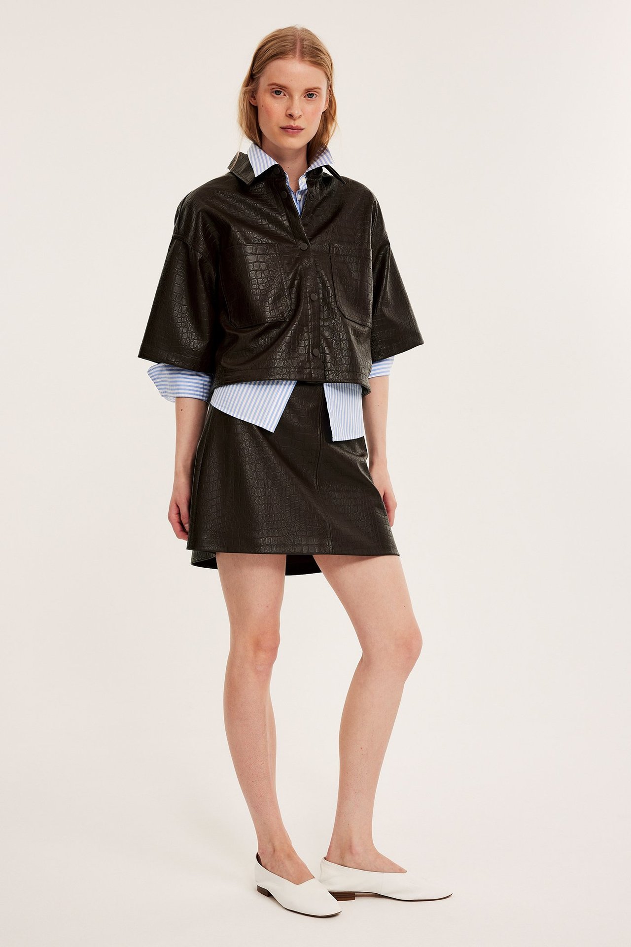 Nainen pukeutuu tummanruskeaan kuvioituun lyhyeen keinonahkapaitaan, raidalliseen paitaan, yhteensopivaan hameeseen, valkoisiin ballerinakenkiin.