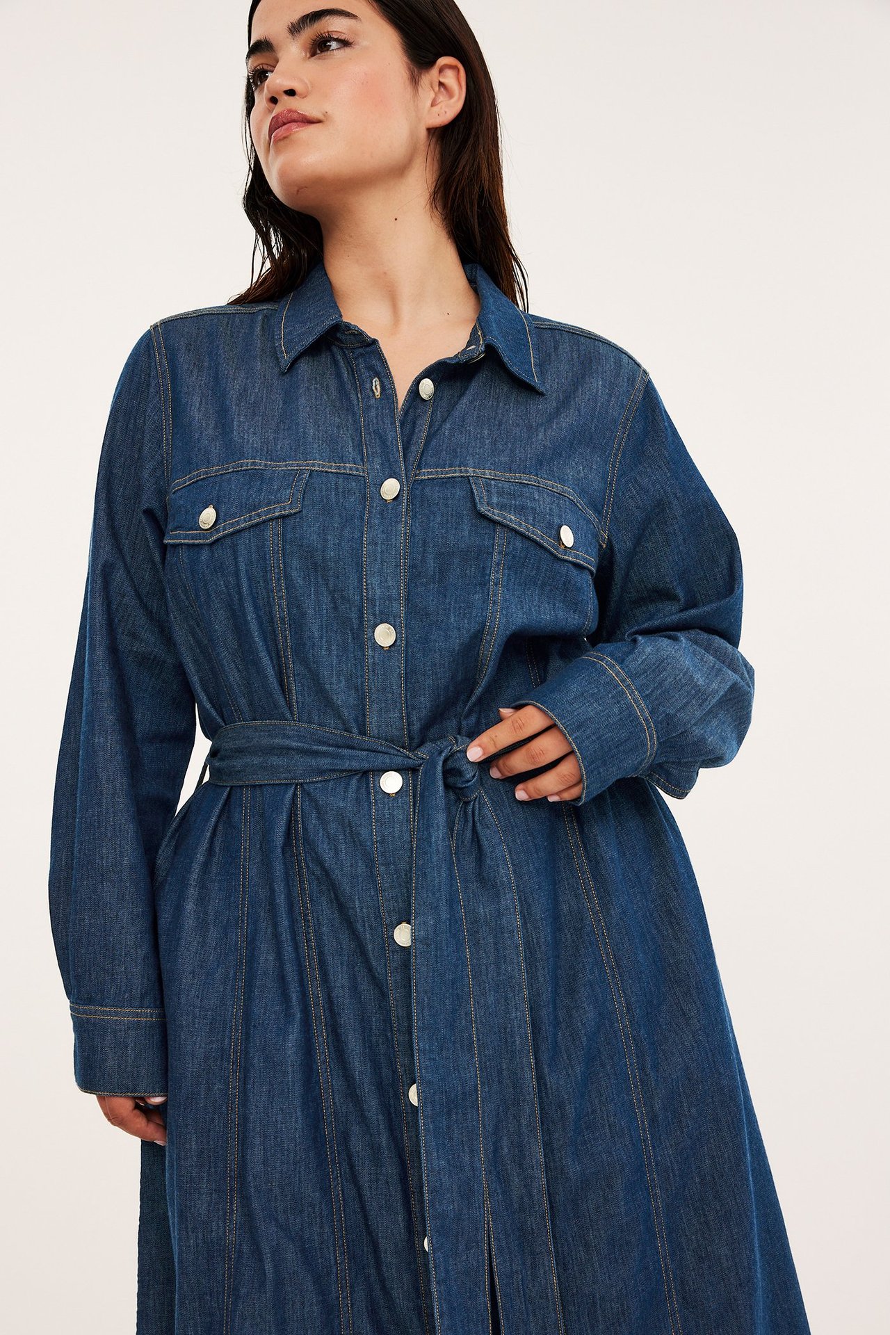 Edestä: nainen tummansinisessä pitkässä denim-mekossa, jossa vyö, rintataskut ja napit.