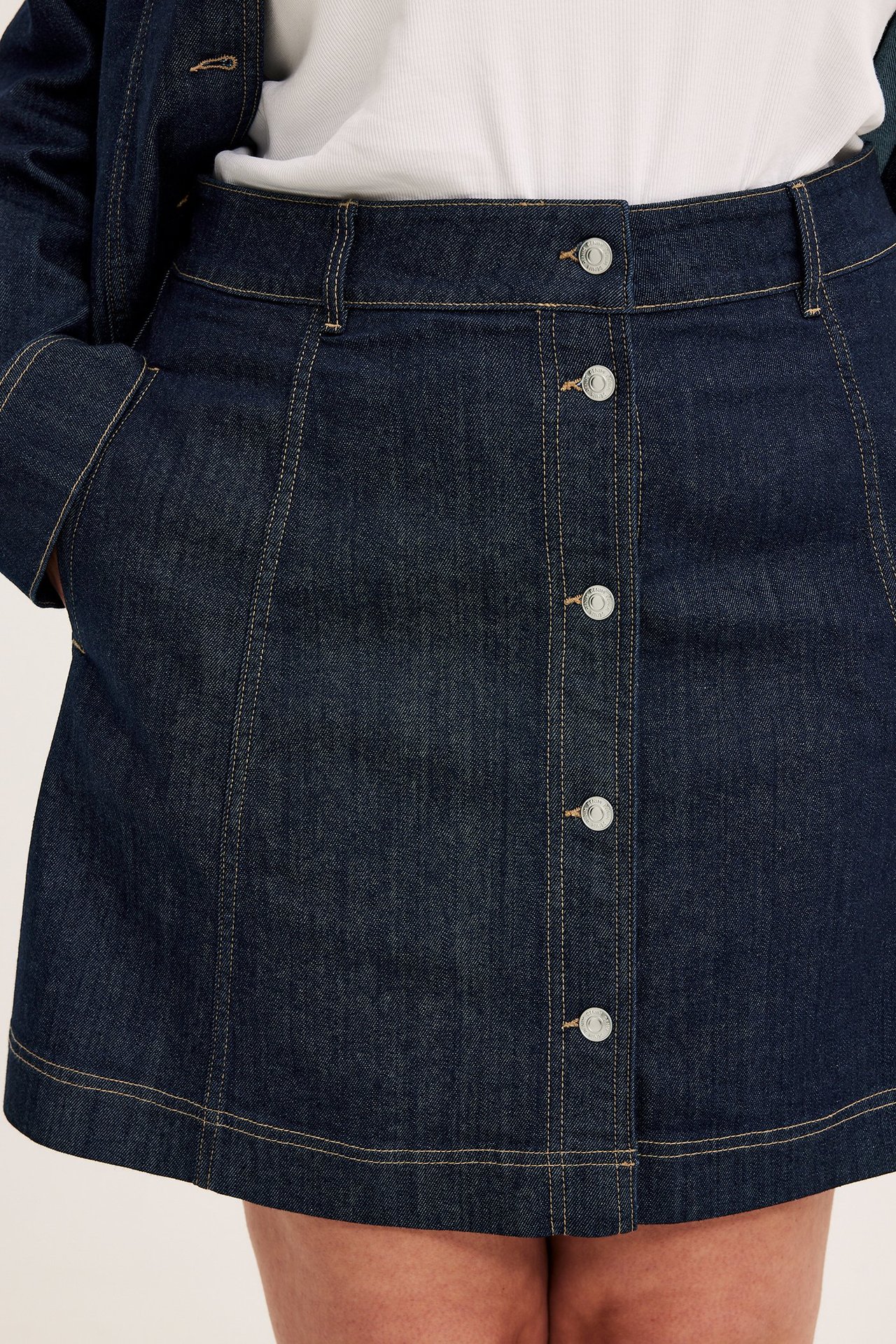 Framifrån: kvinna i mörkblå A-linje jeanskjol med silverknappar, vit topp och jeansjacka.