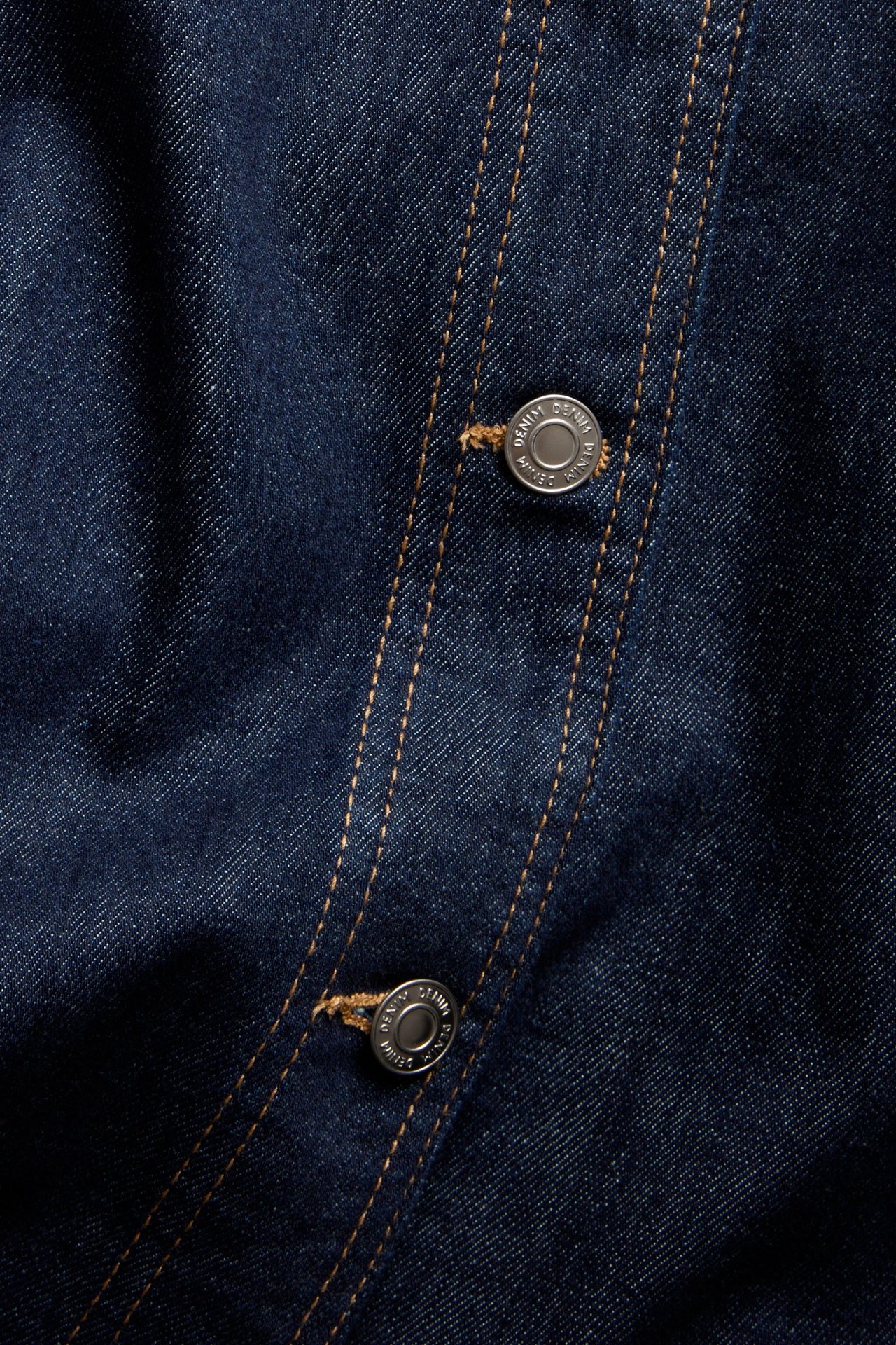 Zbliżenie na ciemnoniebieską spódnicę jeansową w kształcie litery A, ze srebrnymi guzikami i pomarańczowymi szwami.