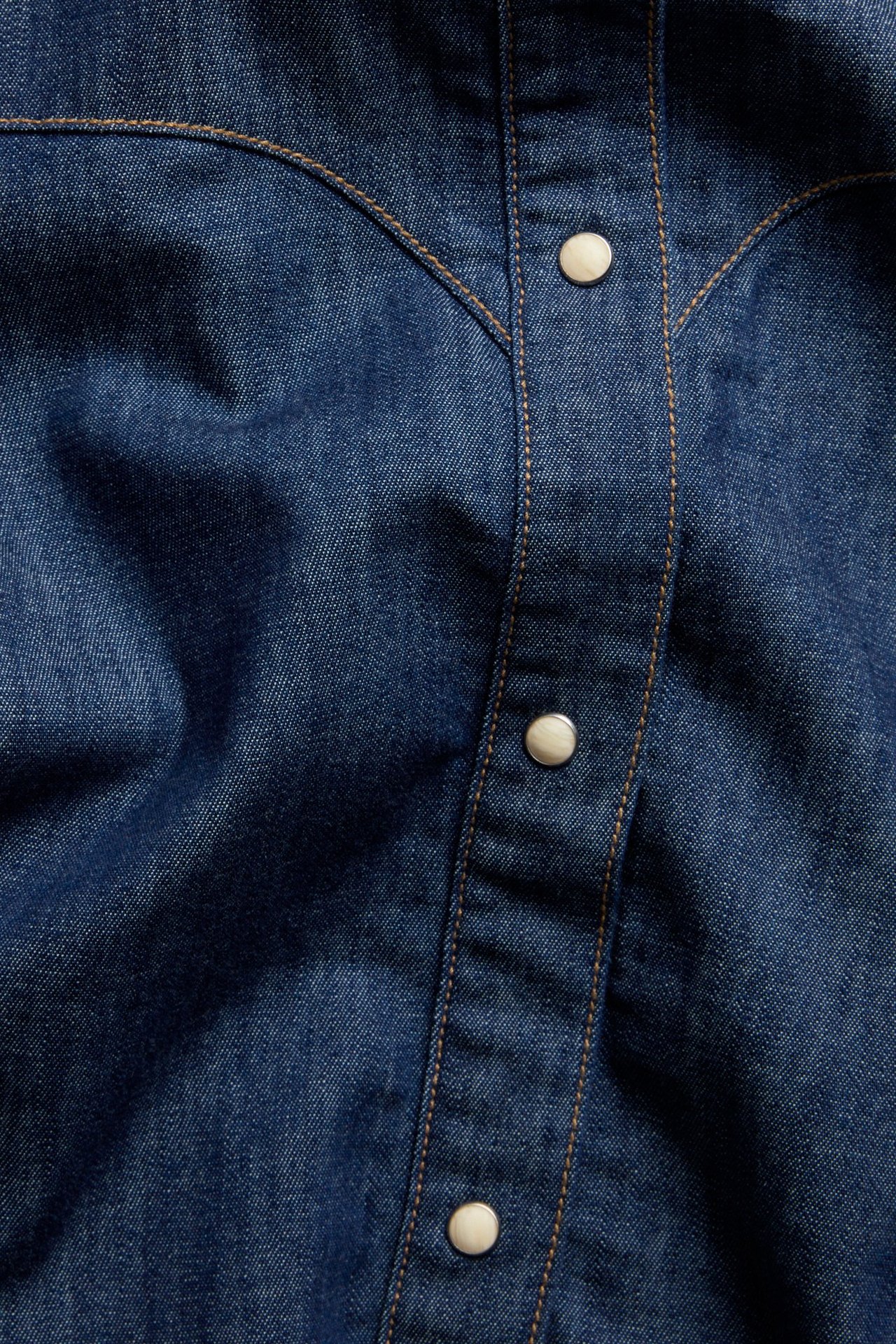 Zbliżenie niebieskiej koszuli jeansowej z karczkiem w stylu western i guzikami zatrzaskowymi w kolorze ecru.