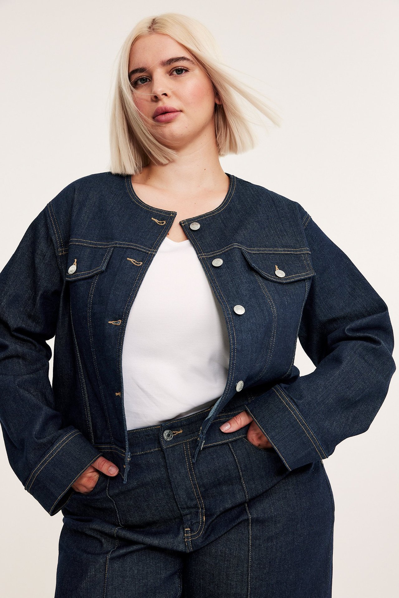 Framifrån: kvinna i mörkblå kort jeansjacka, vit topp och matchande mörkblå jeansbyxor.