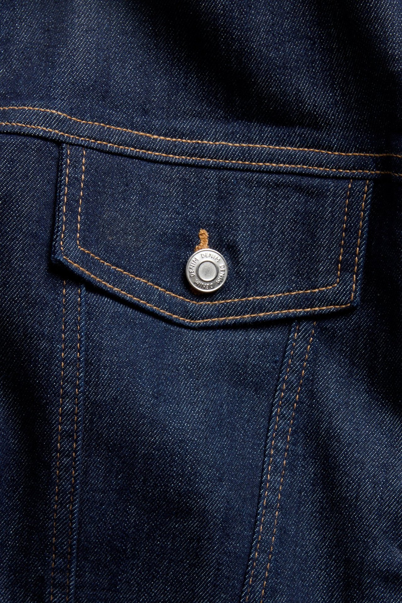 Zbliżenie na ciemnoniebieską kurtkę jeansową z kieszenią z klapką i srebrnym guzikiem, z brązowymi szwami.