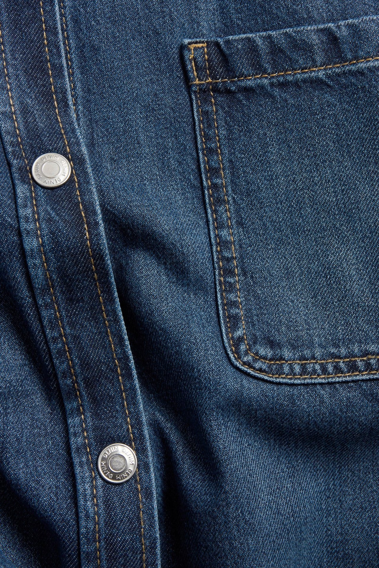 Zbliżenie na oversize'ową niebieską koszulę jeansową ze srebrnymi zatrzaskami i kieszenią na piersi z widocznymi szwami.