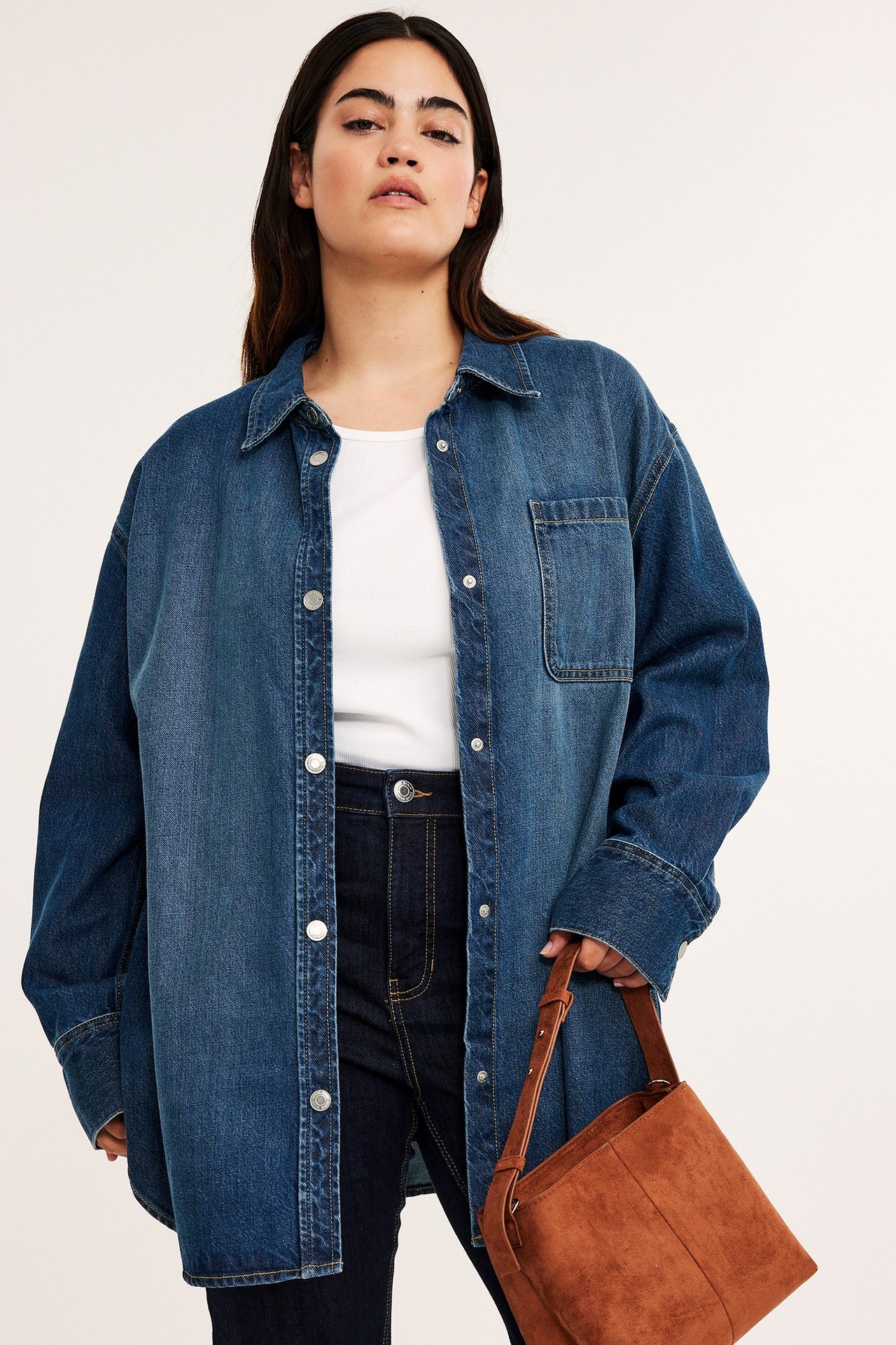 Edestä: Nainen yllään ylisuuri sininen farkkupaita painonapeilla, valkoinen toppi, tummat farkut, ruskea laukku.