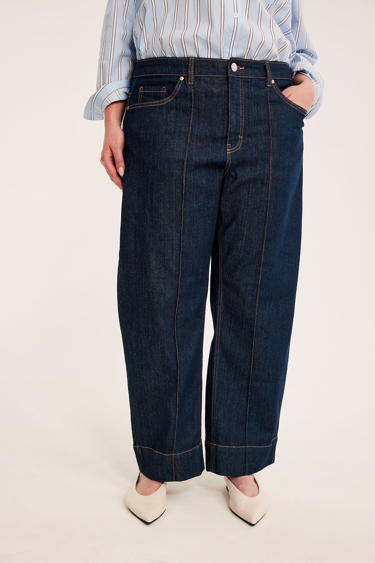 Framifrån, en kvinna i mörkblå vida jeans, ljusblå randig skjorta och beigefärgade spetsiga skor.
