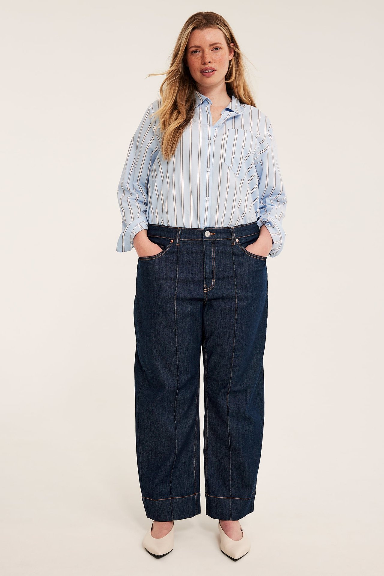 Framsida: kvinna i mörkblå vida jeans, en blårandig skjorta och ljusbeige klackskor.