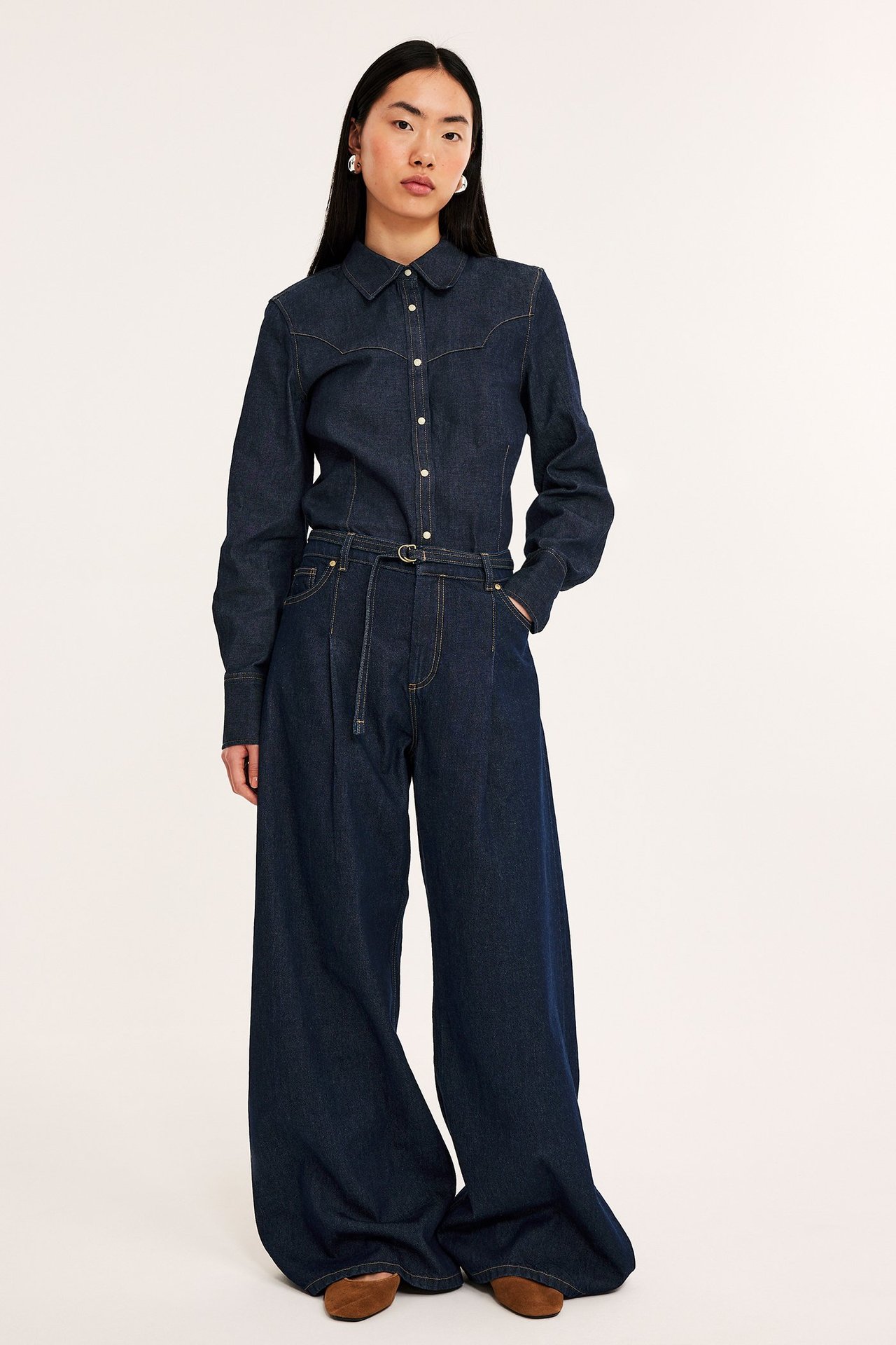 Framifrån, kvinna i figursydd mörkblå jeansskjorta, vida jeans och bruna platta skor.