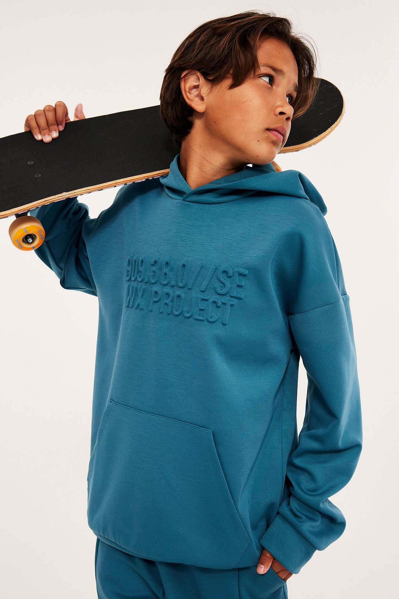 Widok z przodu: Dziecko w zielonej, obszernej bluzie z kapturem z interlocku z nadrukiem 3D, trzyma deskorolkę.
