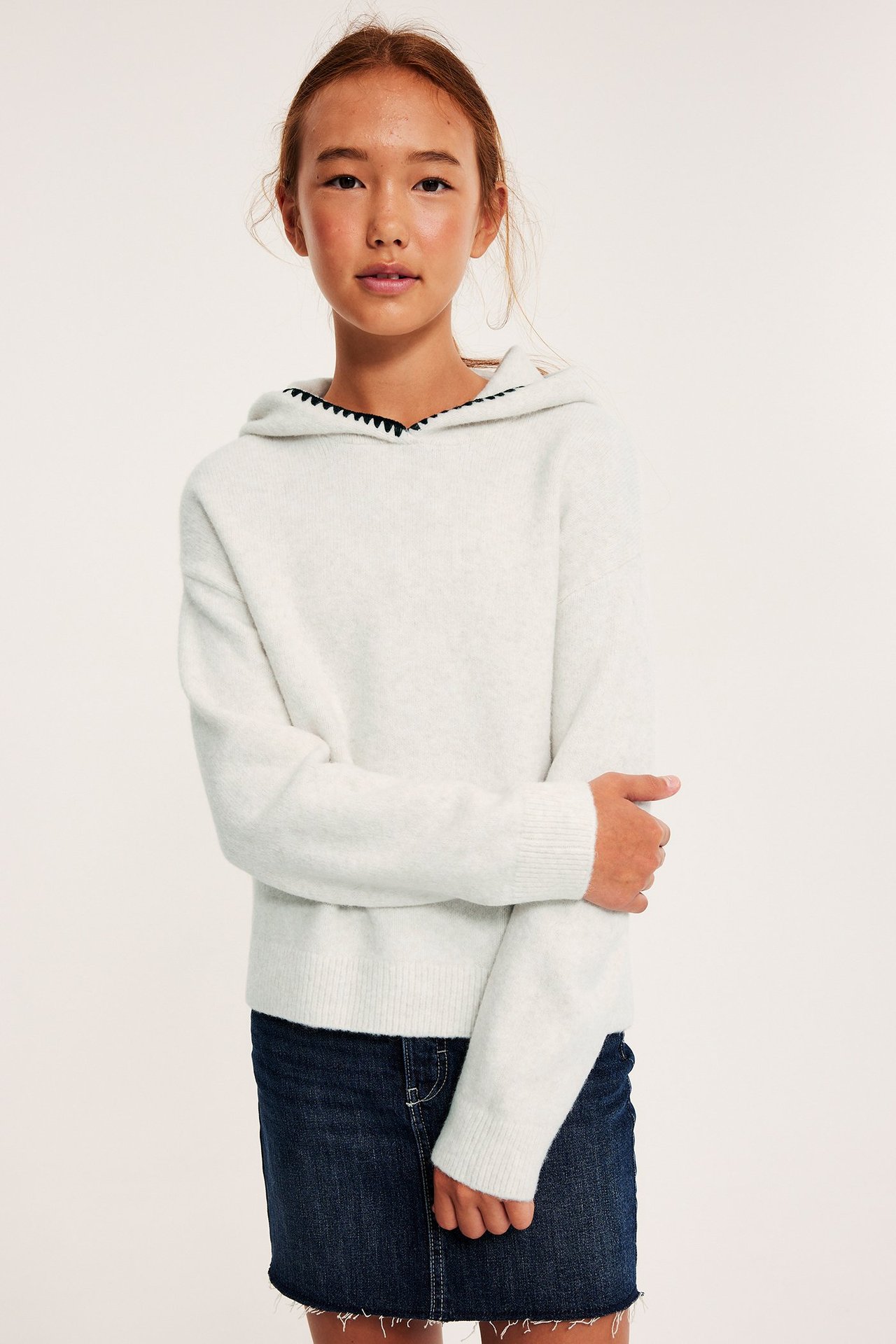 Edestäpäin: Lapsi valkoisessa meleeratussa neulehupparissa mustilla tikkauksilla ja tummansinisessä farkkuhameessa.