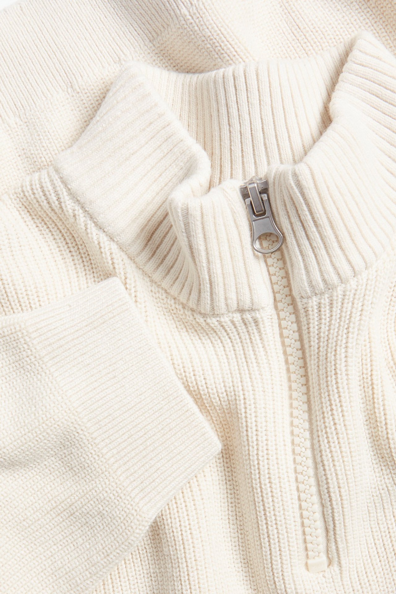 Zbliżenie na jasnobeżowy dzianinowy sweter bawełniany z półzamkiem, prążkowanym kołnierzem i mankietami.