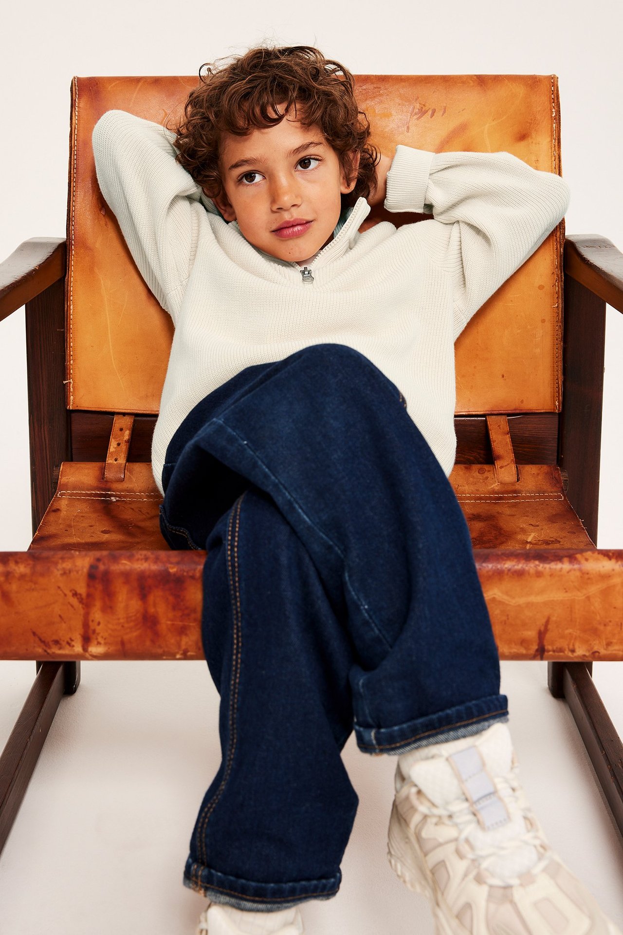 Widok z przodu: dziecko w jasnobeżowym melanżowym bawełnianym swetrze z półzamkiem, ciemnych dżinsach i jasnych sneakersach.