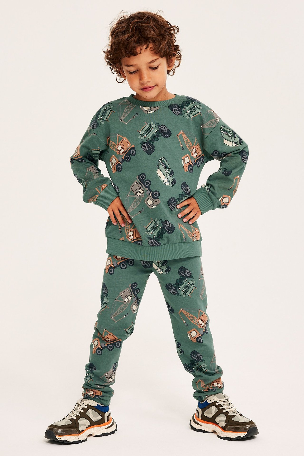 Edestä: lapsi tummanvihreässä puuvillapaidassa ajoneuvoprintillä ja yhteensopivissa collegehousuissa.