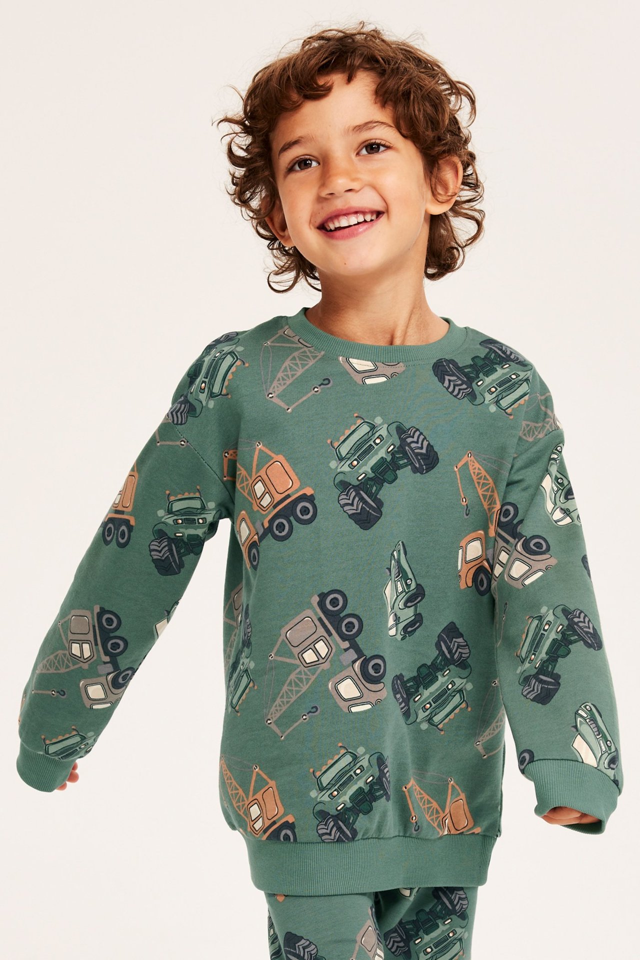 Widok z przodu uśmiechniętego dziecka w ciemnozielonej bawełnianej bluzie z wzorem pojazdów budowlanych.