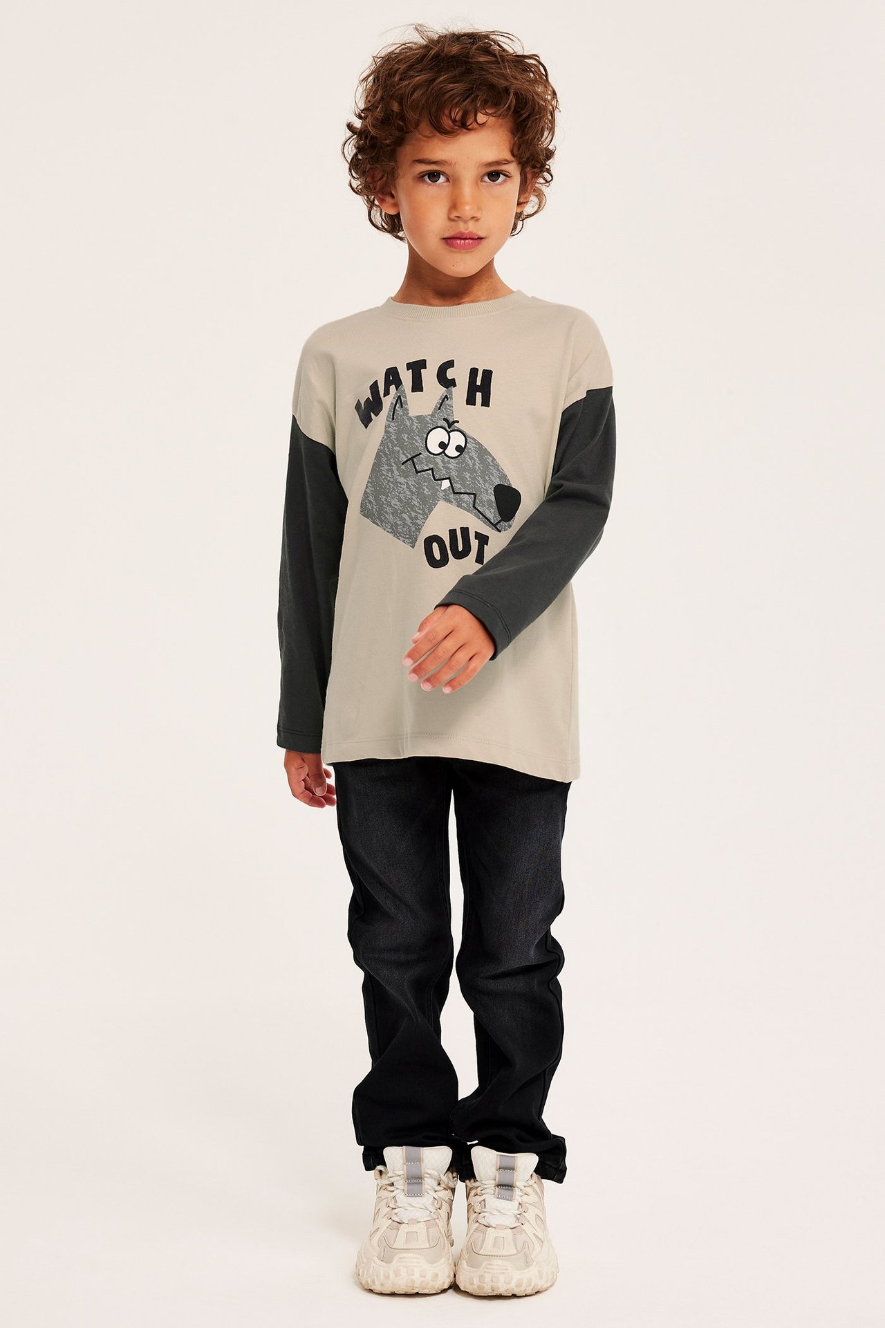 Framifrån: Ett barn i beige långärmad t-shirt med vargtryck, mörka jeans och beiga sneakers.