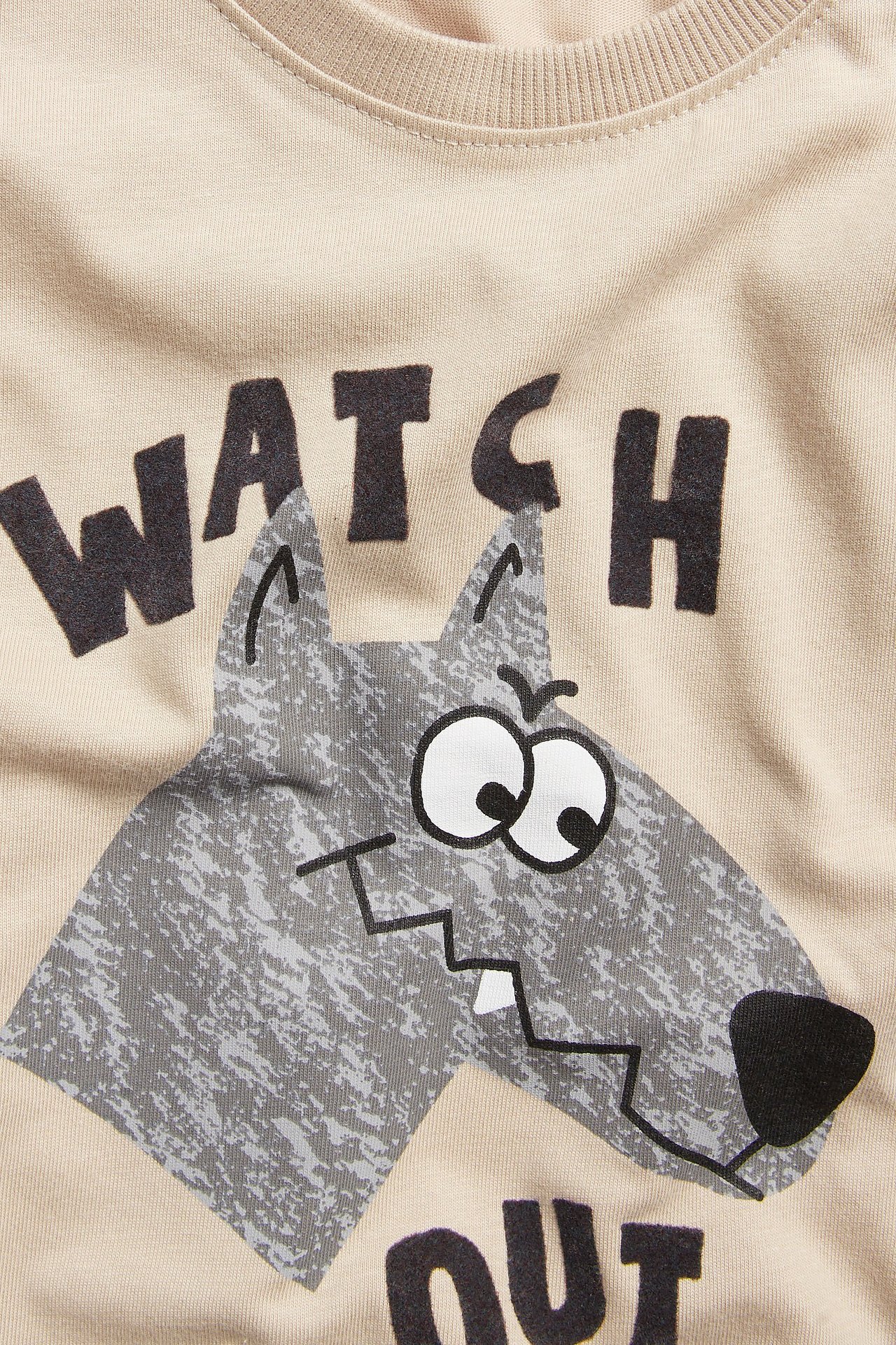 Zbliżenie beżowej bawełnianej koszulki dziecięcej z długim rękawem, z szarym wilkiem i napisem "WATCH OUT".