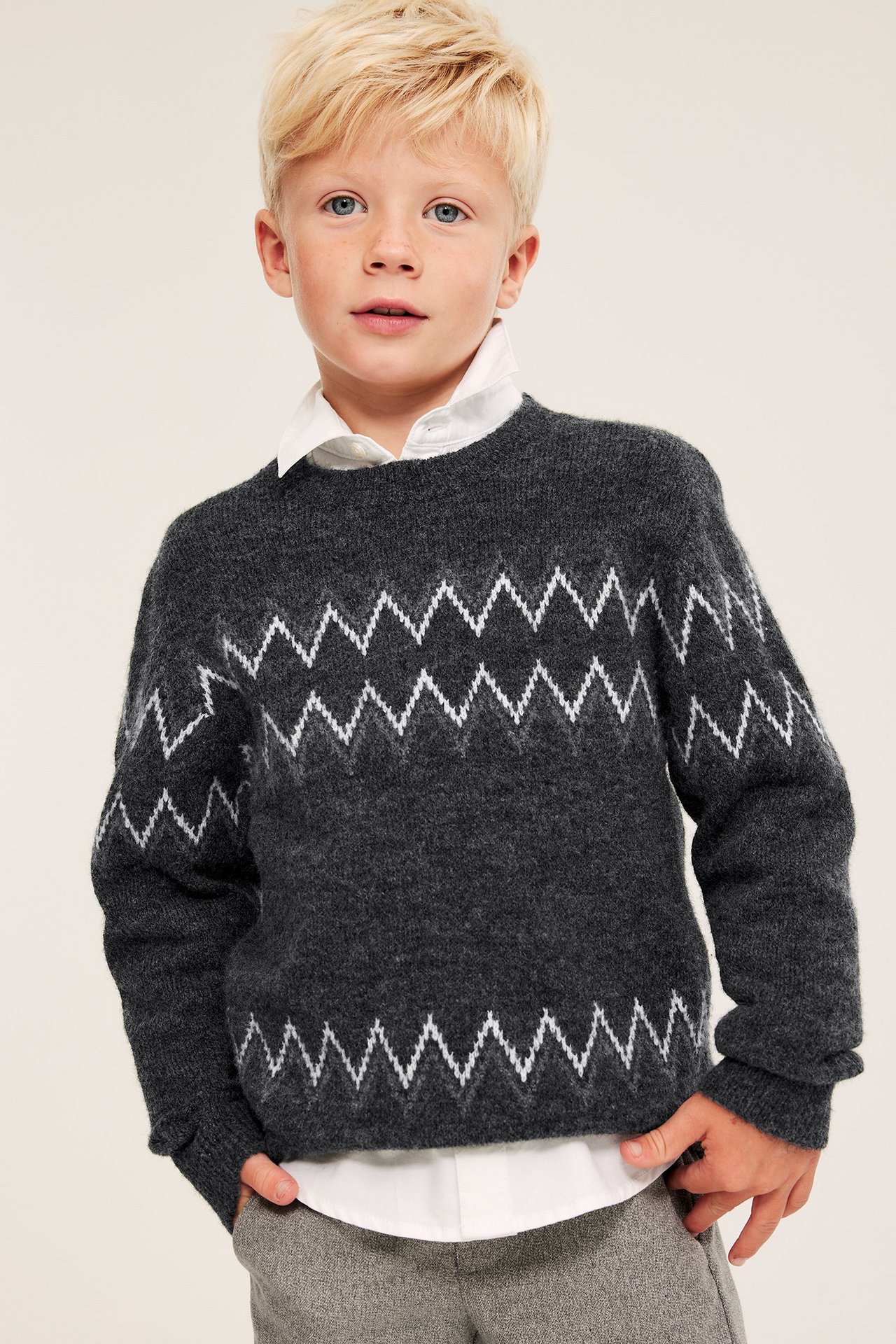 Frontvy av pojke i mörkgrå stickad tröja med vitt sicksackmönster över en vit skjorta.