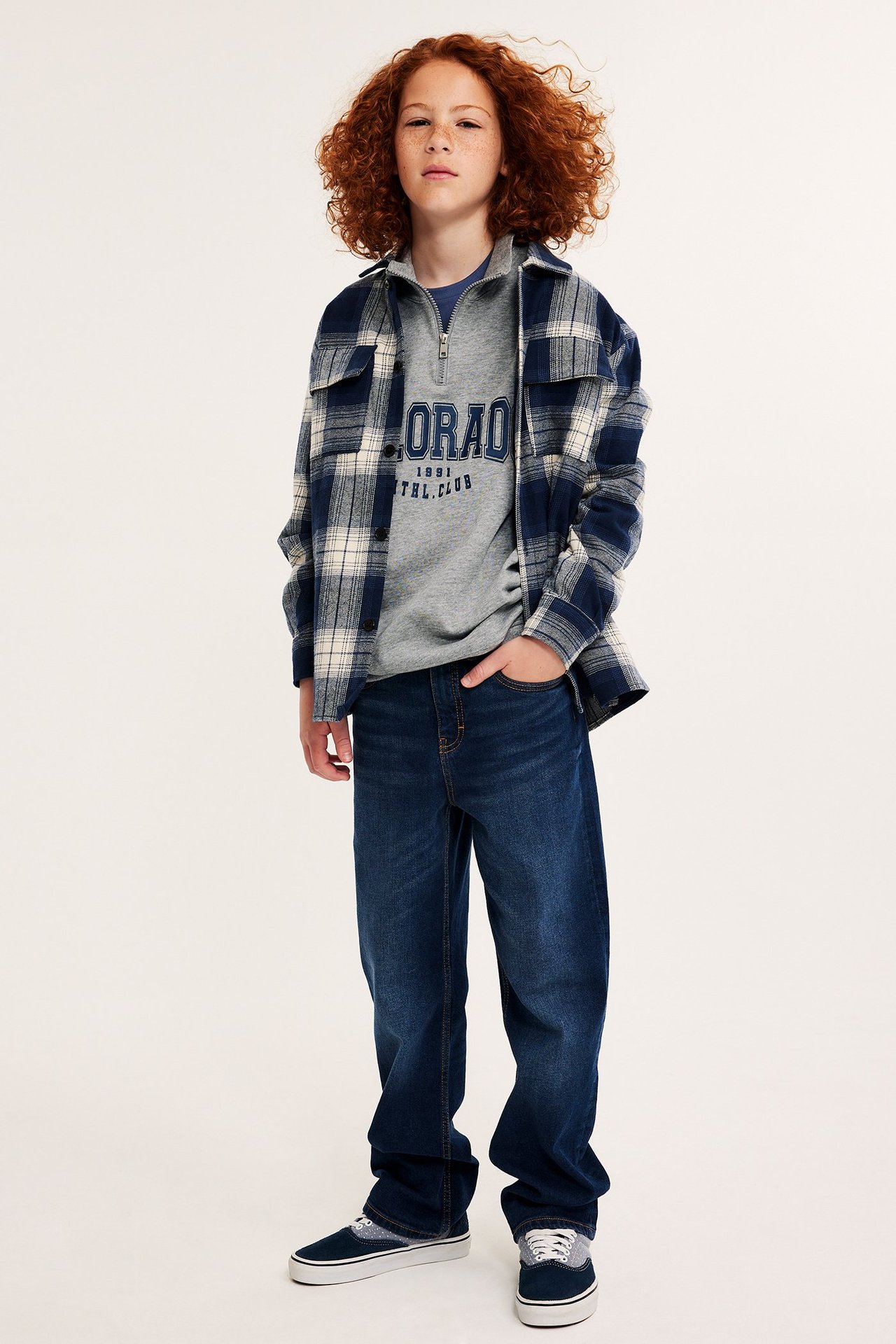 Framsida av ett barn i mörkgrå sweatshirt med dragkedja, blårutig skjorta, jeans och blå sneakers.