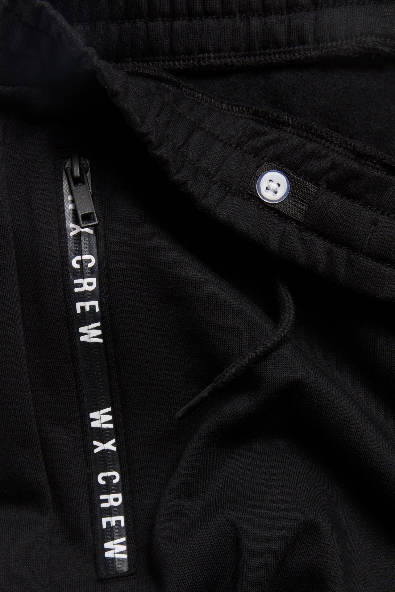 Zbliżenie czarnych spodni dresowych z kieszenią na zamek, nadrukiem 'WX CREW', sznurkiem i guzikiem w pasie.