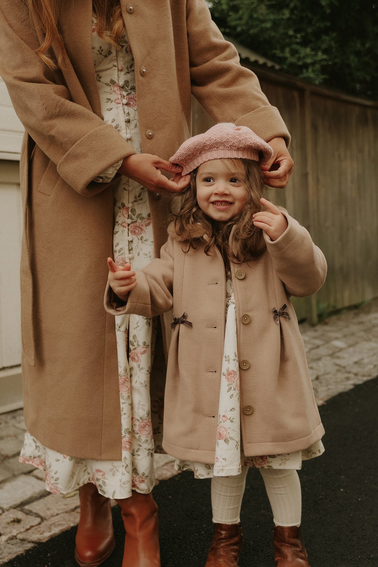 Barn i lysbrun kåpe, blomstret kjole, rosa basker; voksen i lang kamelfarget kåpe.