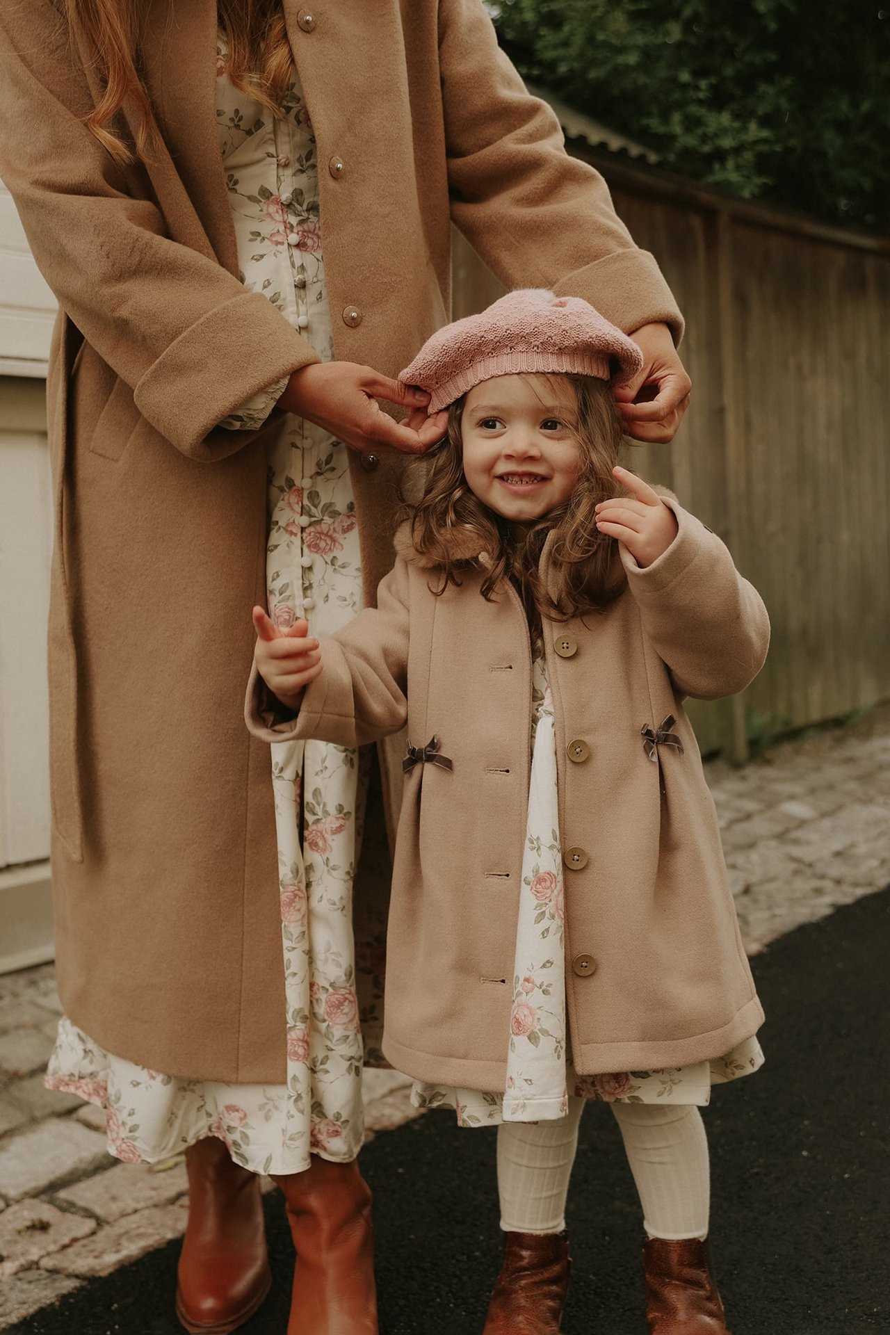 Barn i lysbrun kåpe, blomstret kjole, rosa basker; voksen i lang kamelfarget kåpe.