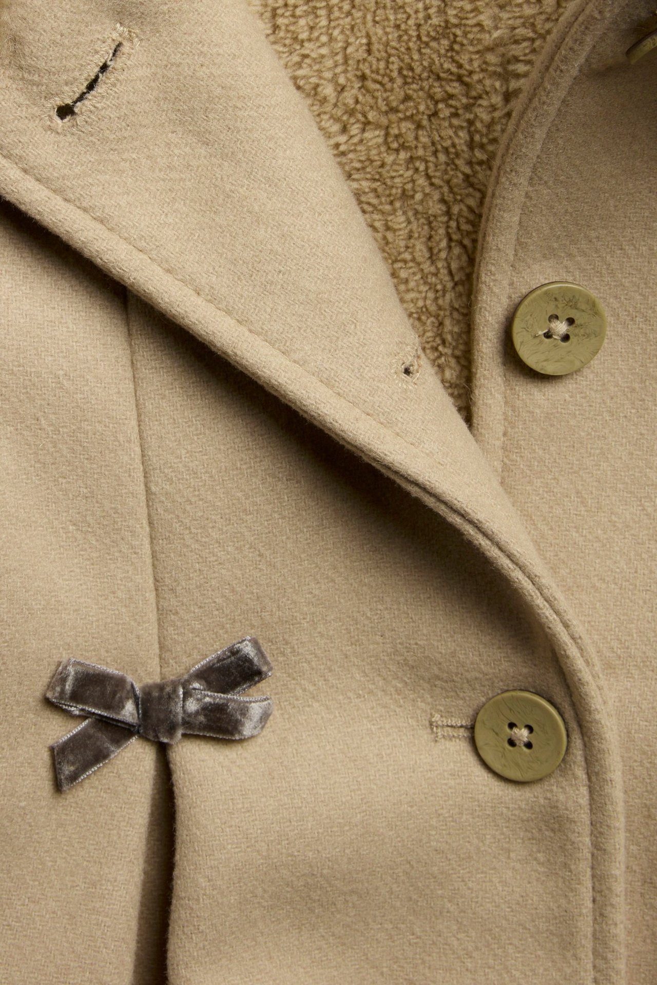 Zbliżenie przodu beżowego płaszcza z wełny, z podszewką, zielonymi guzikami i szarą aksamitną kokardą.