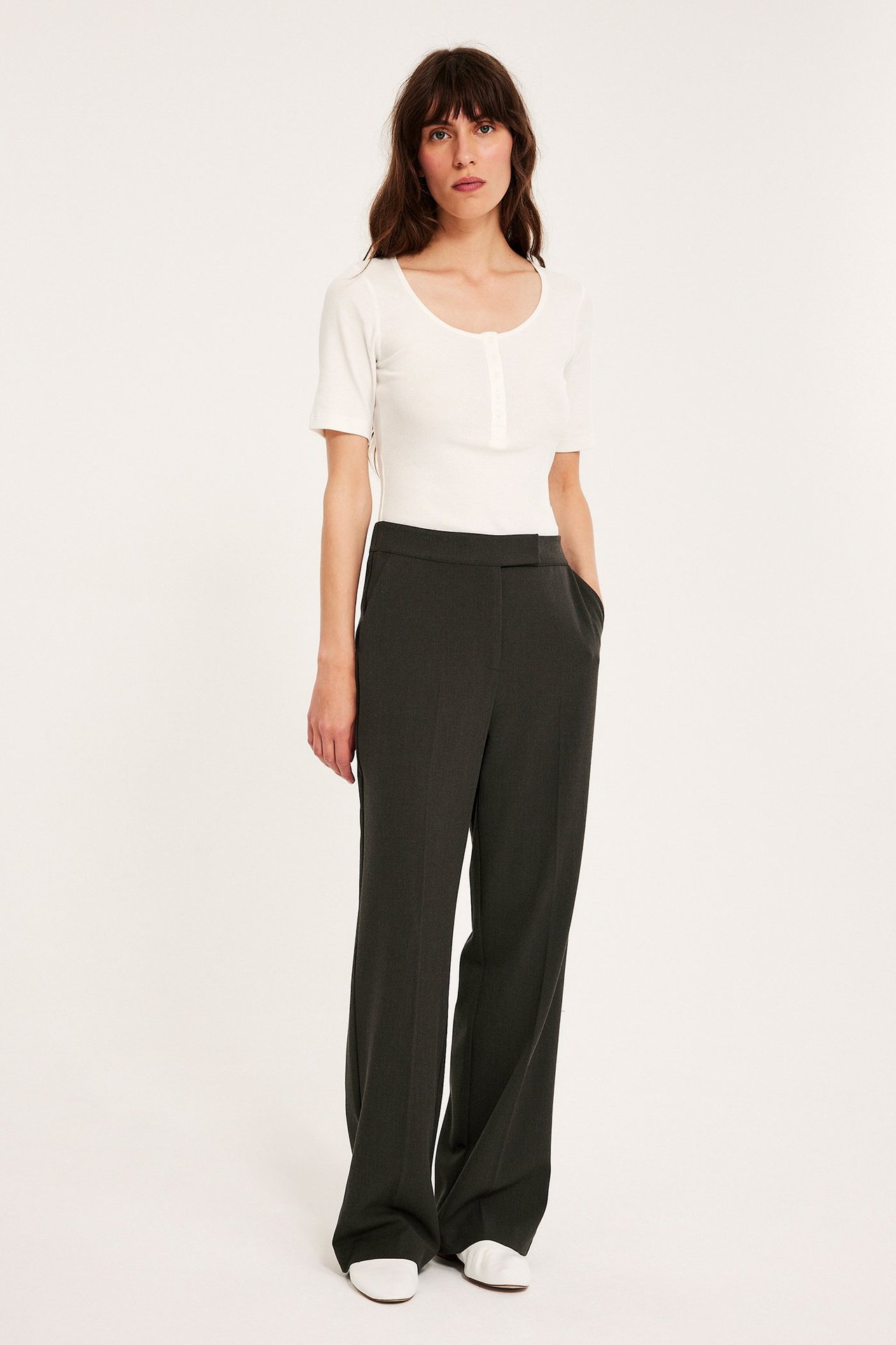 Widok z przodu: kobieta w ciemnobrązowych szerokich spodniach garniturowych, białym prążkowanym topie i białych płaskich butach.
