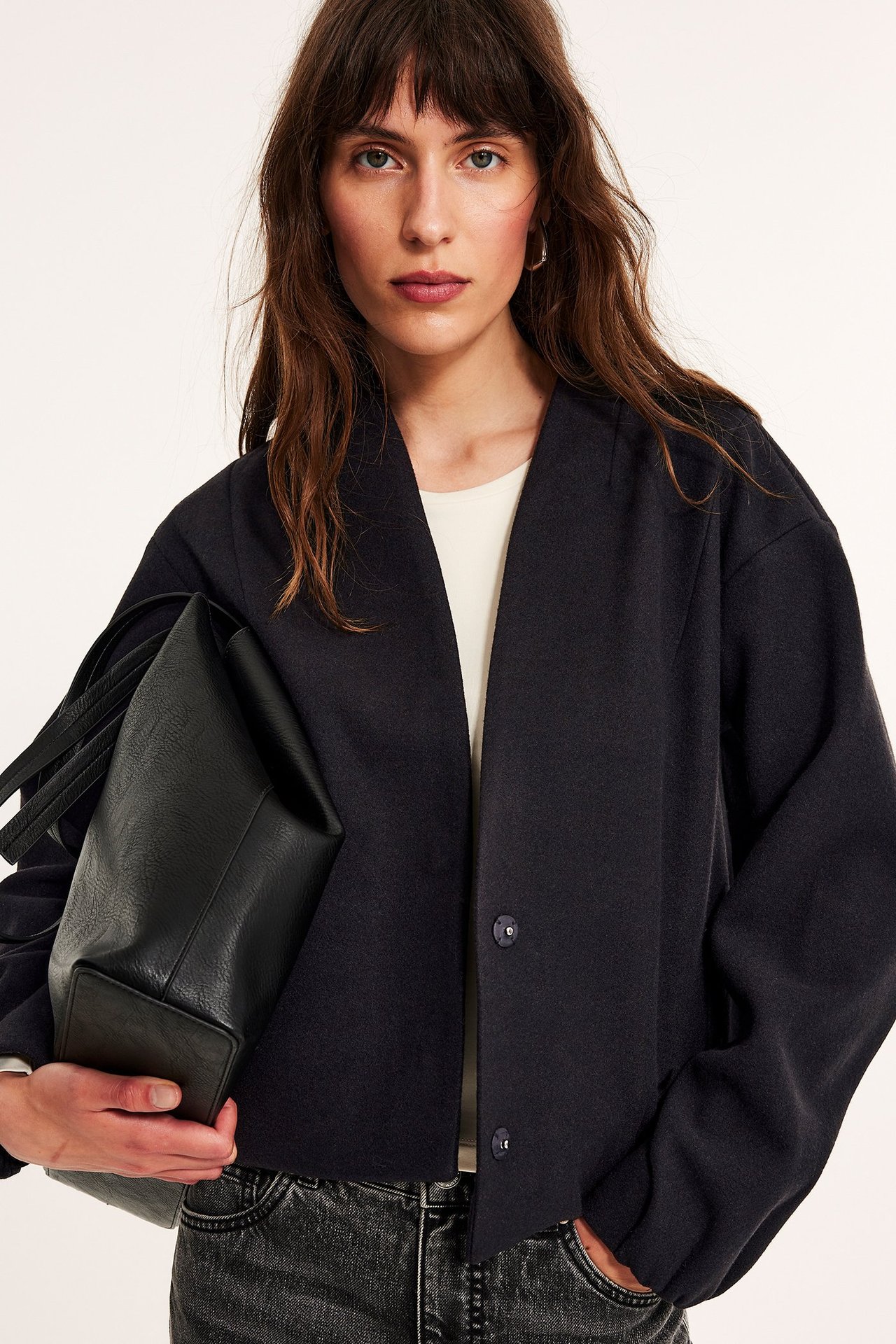 Widok z przodu: Kobieta w czarnej krótkiej kurtce, kremowym topie, czarnych dżinsach, trzyma czarną torebkę.