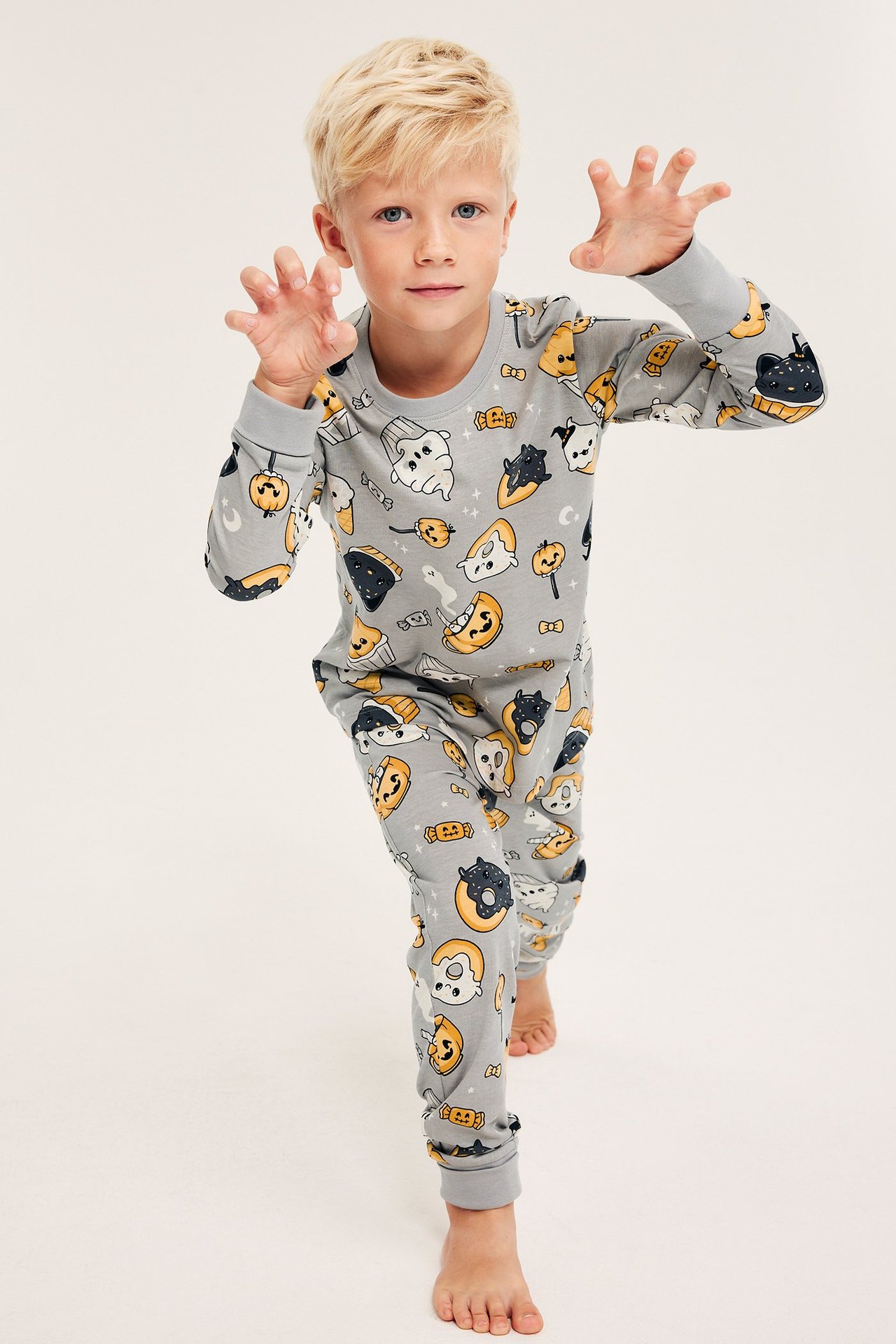 Edestä: lapsi harmaassa puuvillapyjamassa, jossa hauska Halloween-kuvio, tekee pelottavan eleen.