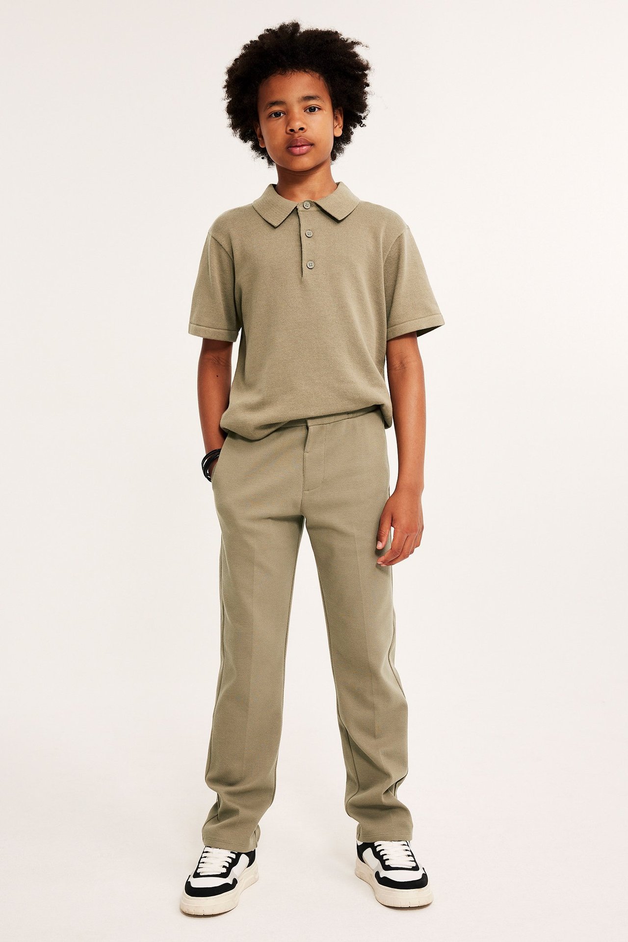 Edestä: lapsi yllään tummanbeiget jerseyhousut prässivekeillä, yhteensopiva pikeepaita ja lenkkarit.