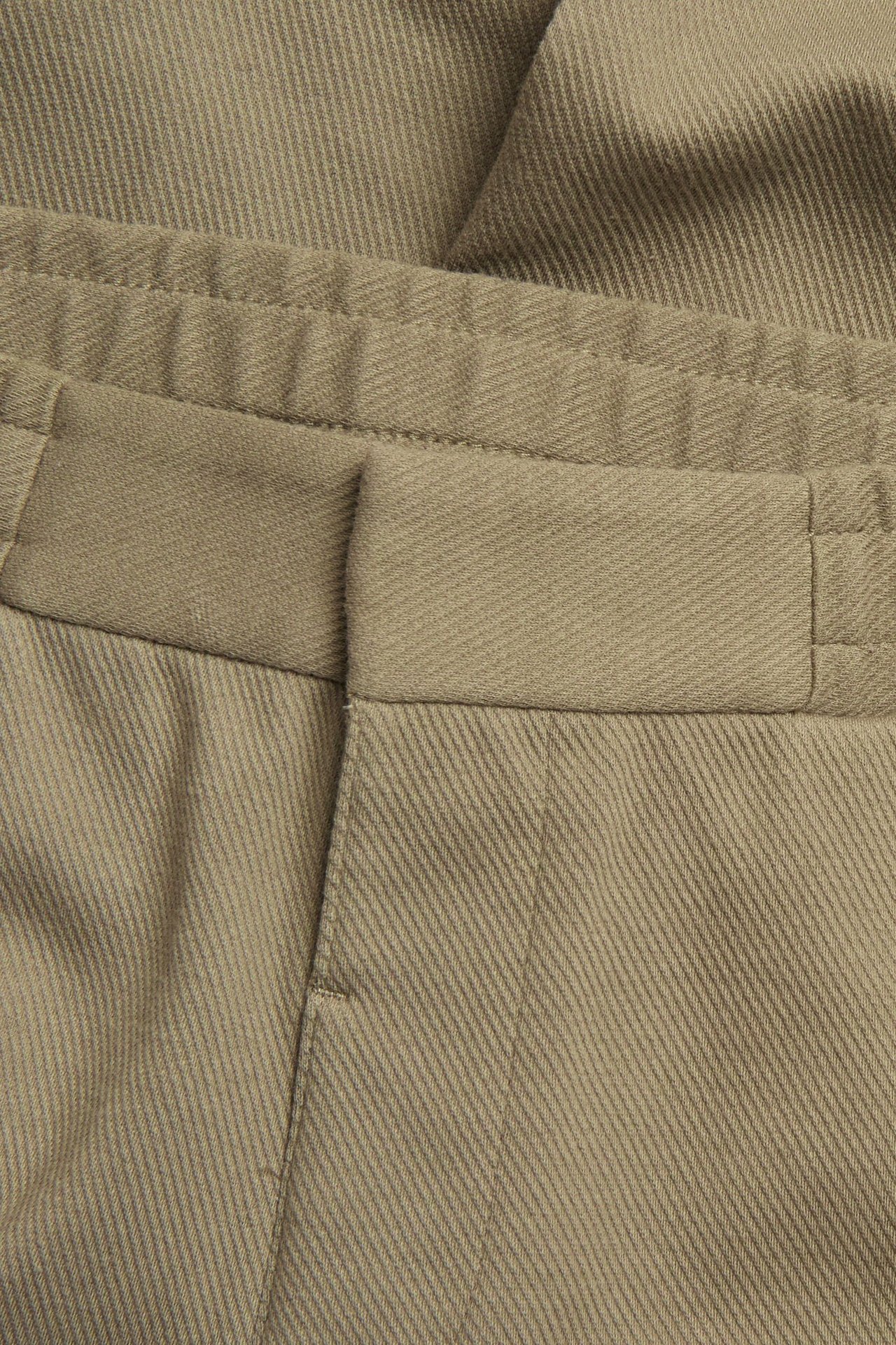 Zbliżenie na ciemnobeżowe spodnie dresowe z kantami, imitacją rozporka i elastyczną talią.