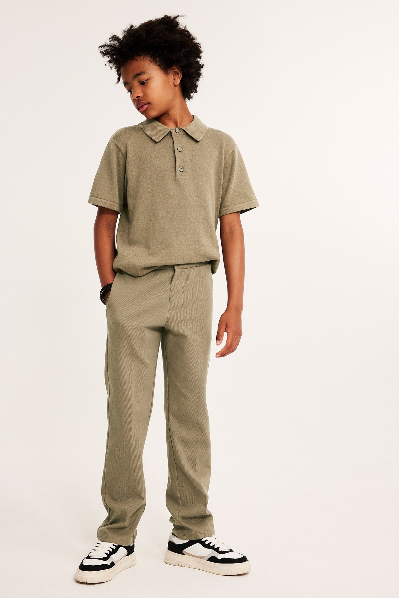 Edestä: Lapsi tummanbeigessä jerseyhousuissa prässivekeillä, yhteensopivassa pikeepaidassa ja lenkkareissa.