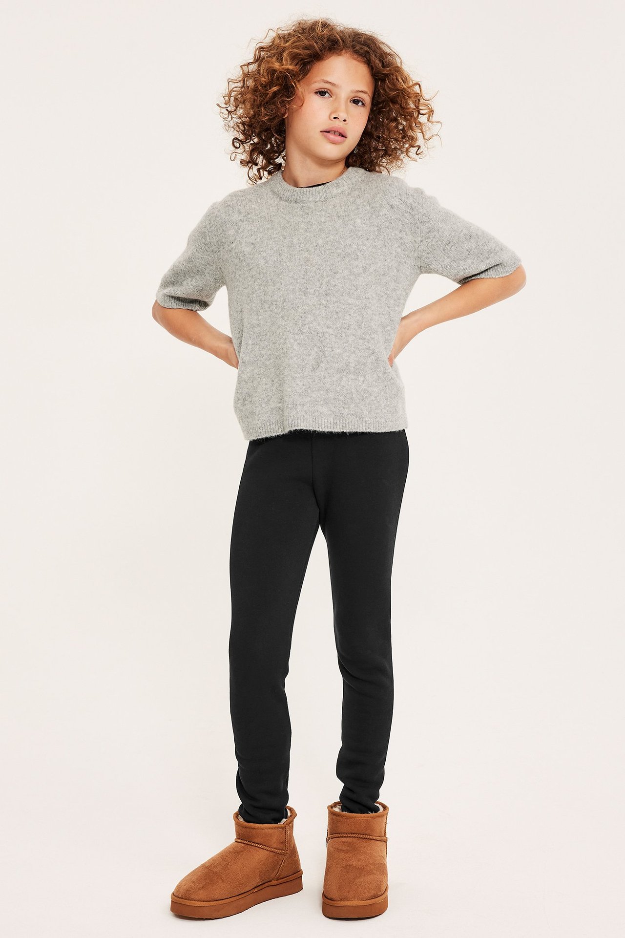 Edestäpäin lapsi mustissa leggingseissä, vaaleanharmaassa lyhythihaisessa neuleessa ja ruskeissa nilkkureissa.