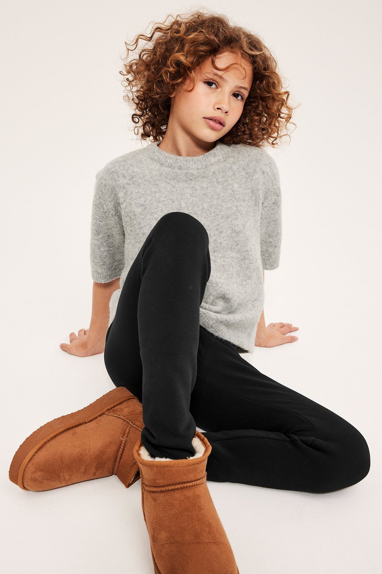 Edestä, istuva lapsi mustissa lämpimissä leggingseissä, vaaleanharmaassa neuleessa ja ruskeissa nilkkureissa.