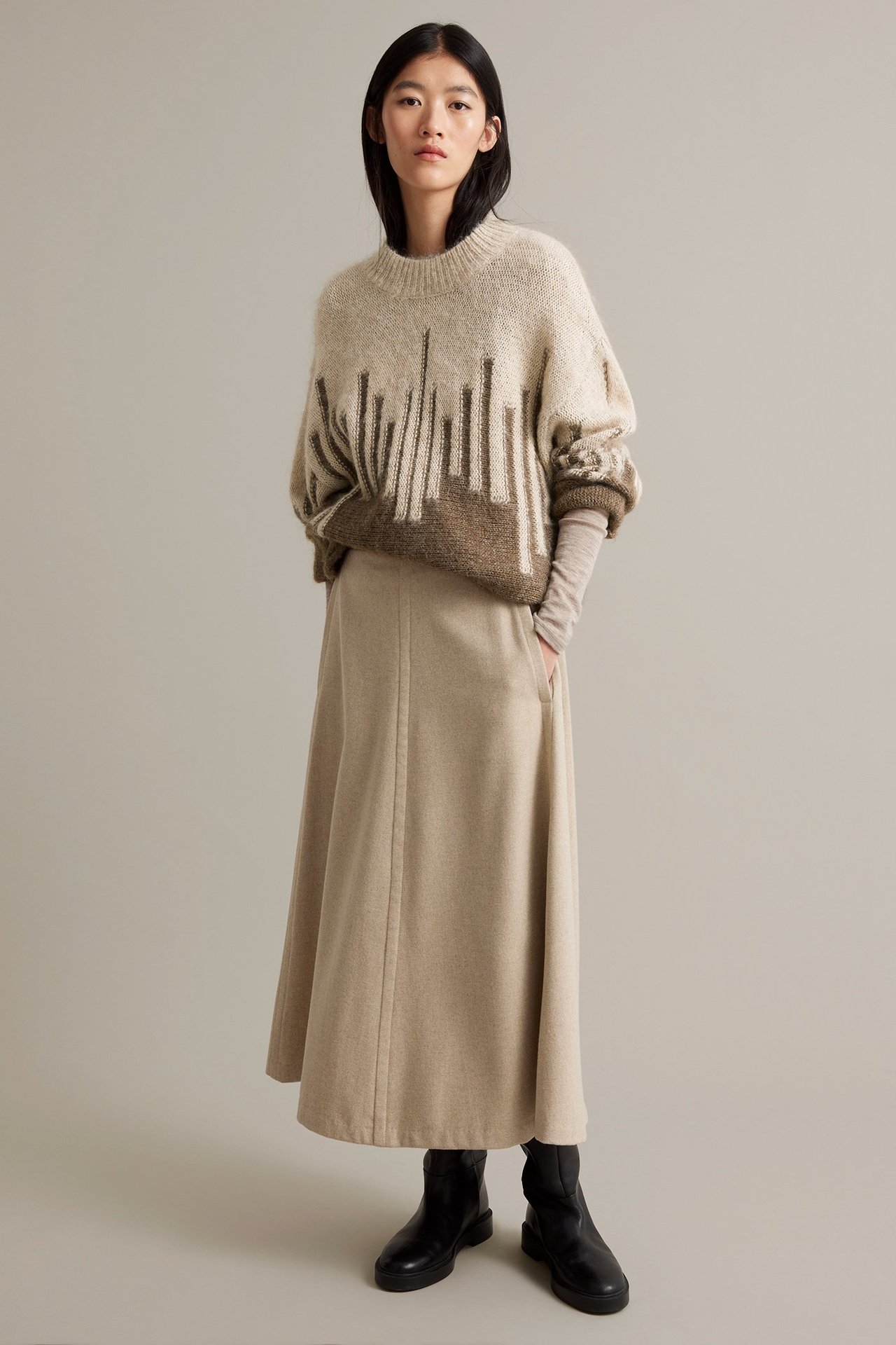 Naismallinen malli beige pitkässä hameessa ja ruskeassa kirjoneuleessa, edestä.