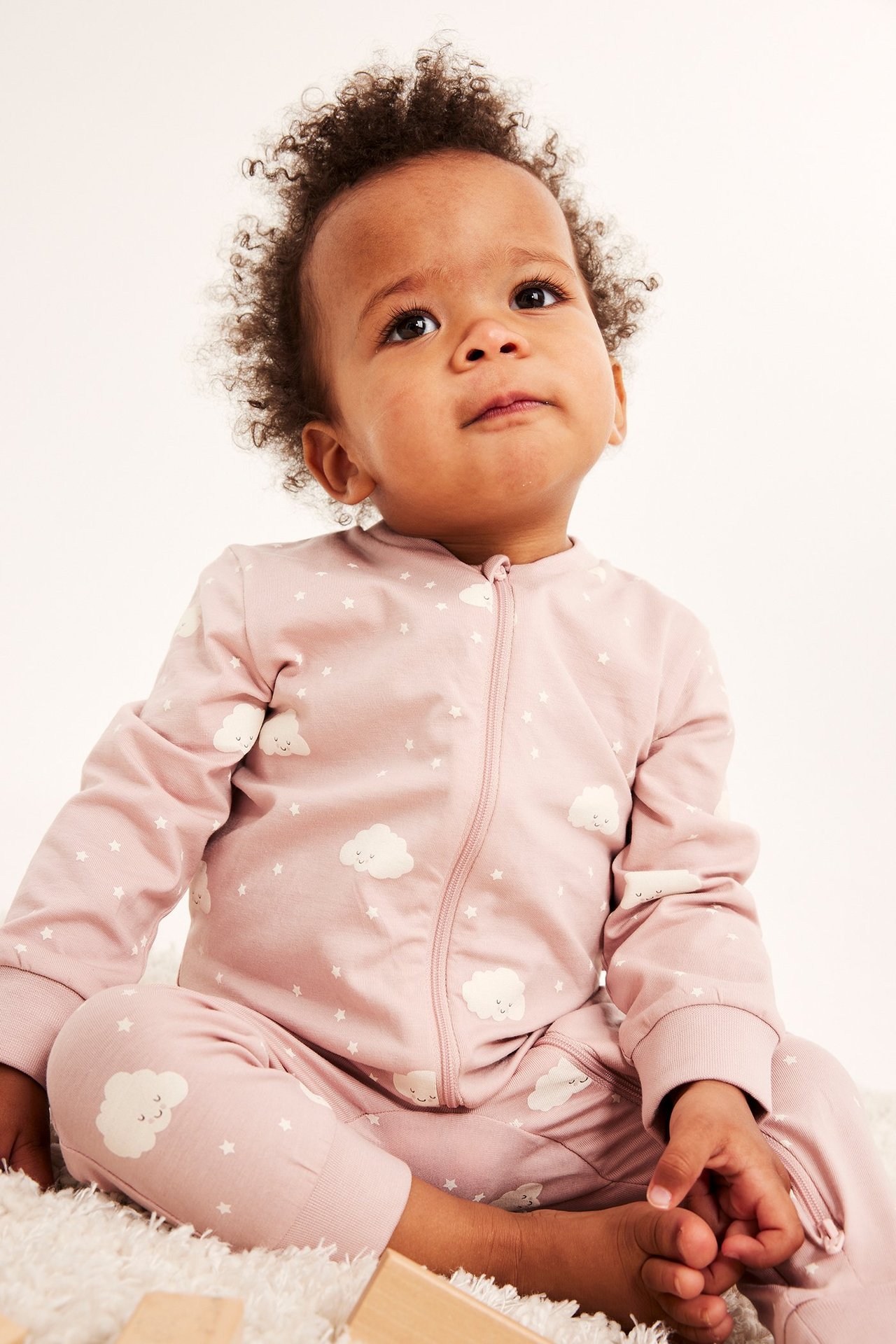 Edestä: Vauva vaaleanpunaisessa kuvioidussa pitkähihaisessa potkupuvussa, jossa valkoisia pilviä ja tähtiä, kaksisuuntainen vetoketju.