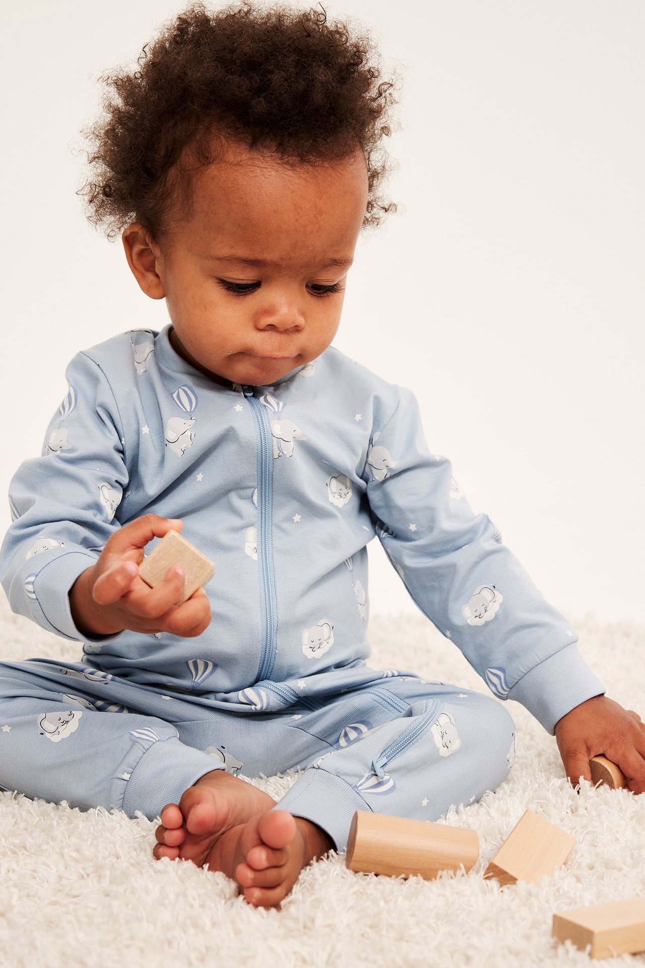 Edestä: vauva vaaleansinisessä kuviollisessa puuvillaromperissa kaksisuuntaisella vetoketjulla, pitää puupalikkaa.