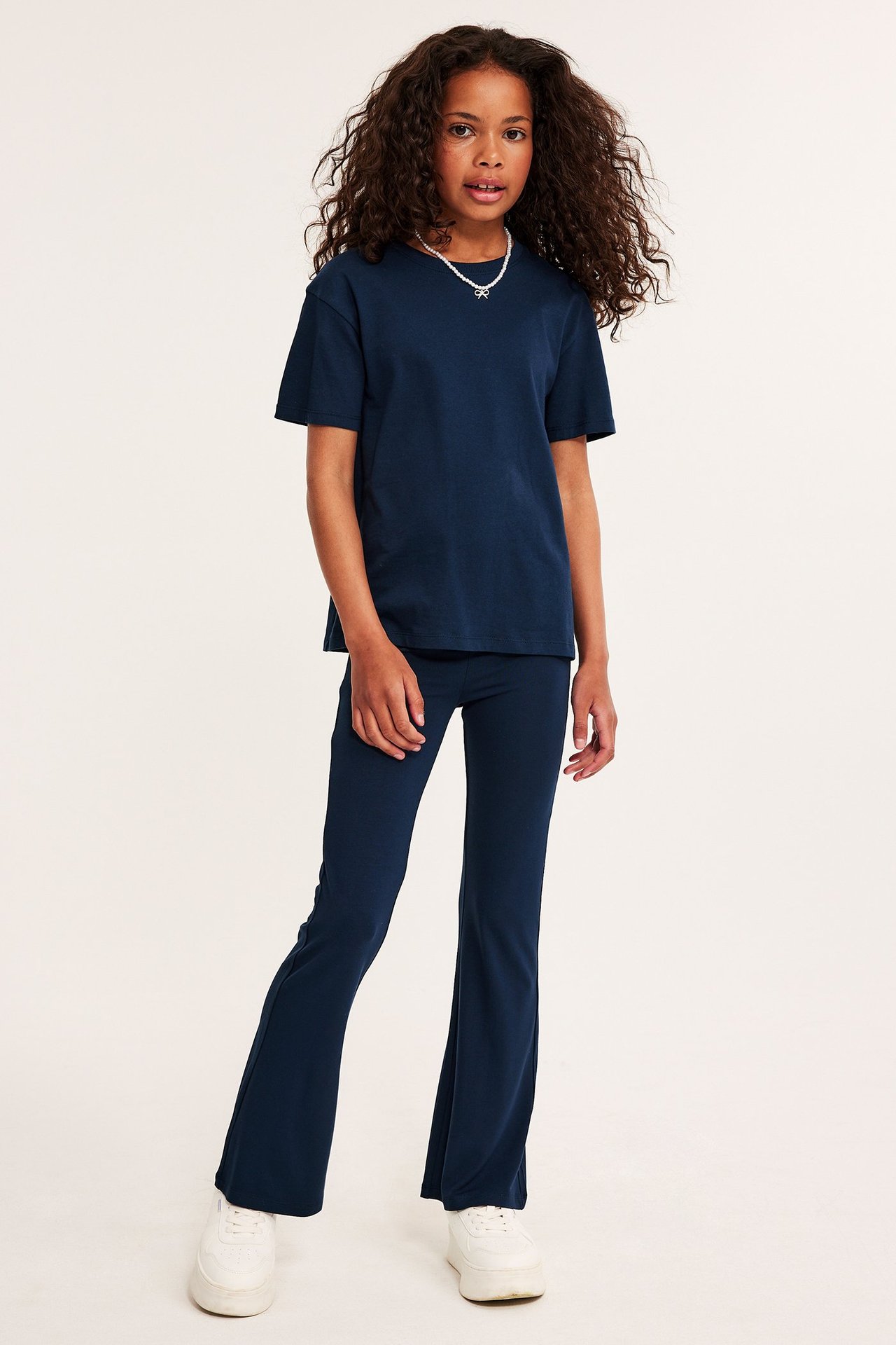 Edestä: lapsi tummansinisissä viskoosi-flare-joogaleggingsseissä, yhteensopivassa t-paidassa, valkoisissa lenkkareissa ja kaulakorussa.